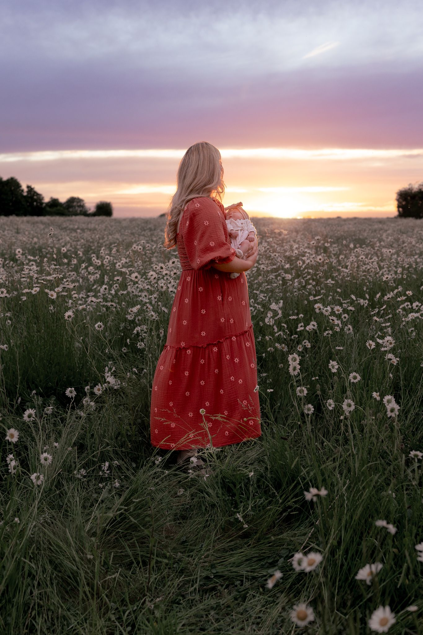 A blonde woman in a red floral dress stands in a wildflower field holding a baby at sunset.