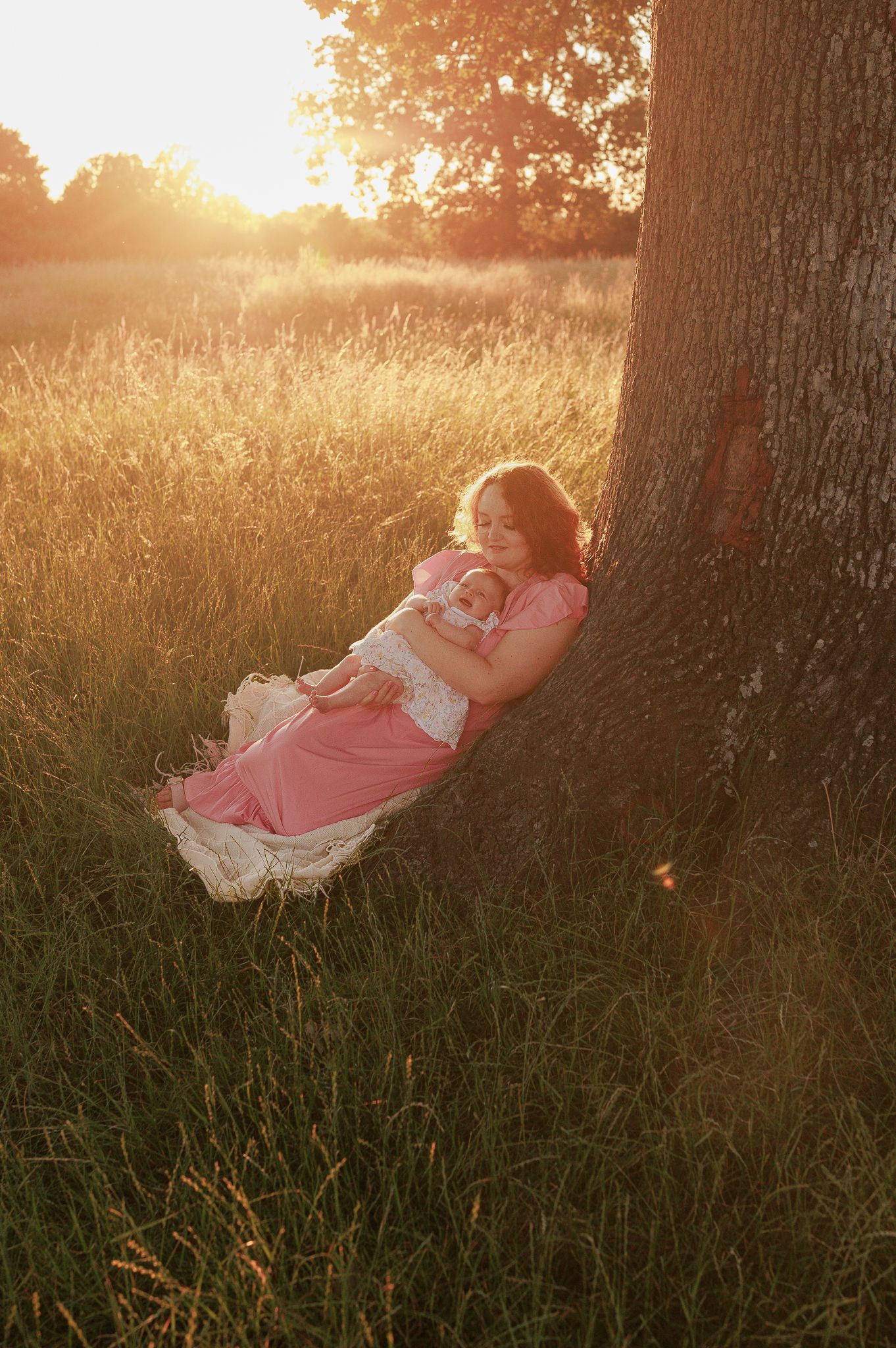 Woman in pink dress cradling newborn baby leaning against tree in golden sunset meadow in Swindon.