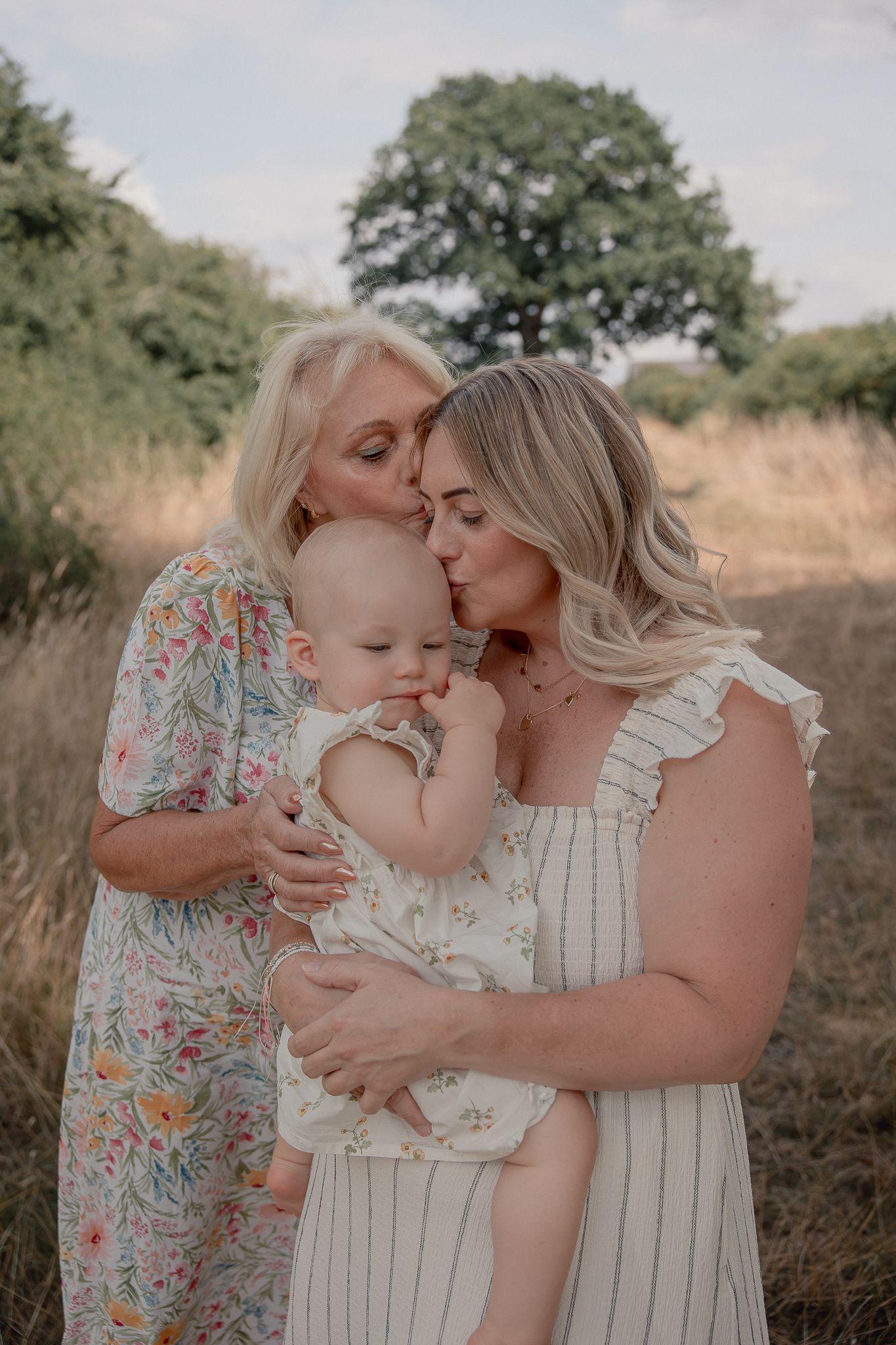 Two women in floral dresses kiss a baby outdoors in a sunny field, capturing a tender three-generation family moment.