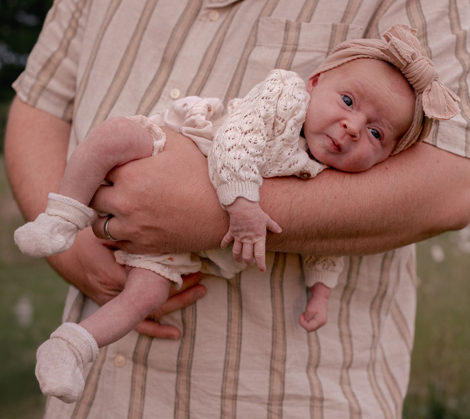 Newborn baby in white knit outfit and bow headband cradled in parent's arms outdoors.