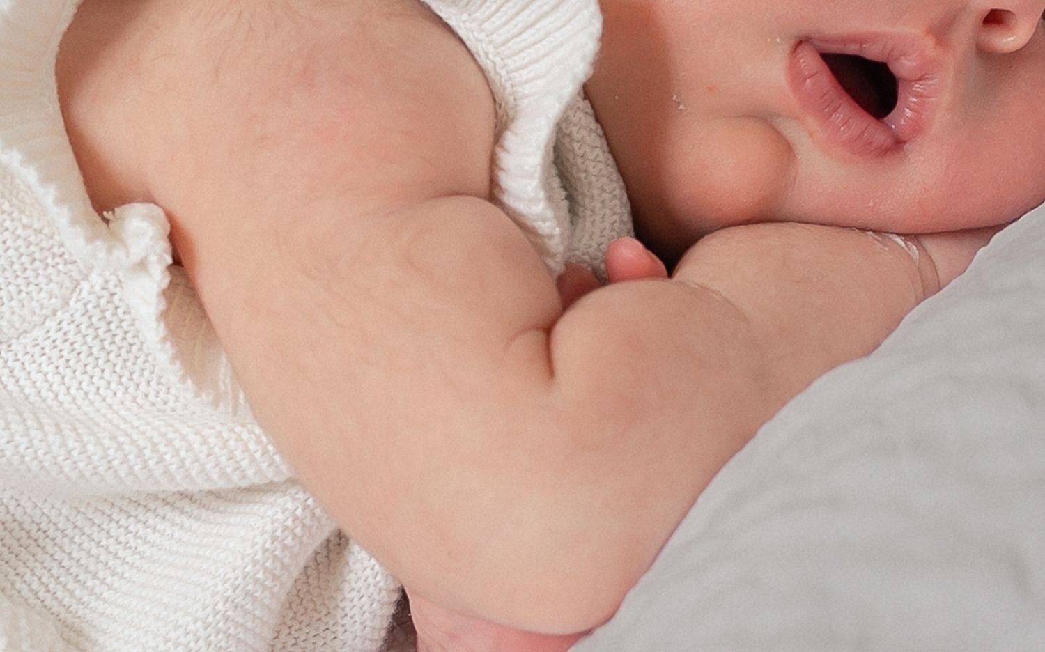 Adorable yawning newborn baby lying on a soft white blanket, showing chubby arms and rosy cheeks in Cirencester, Cotswolds.