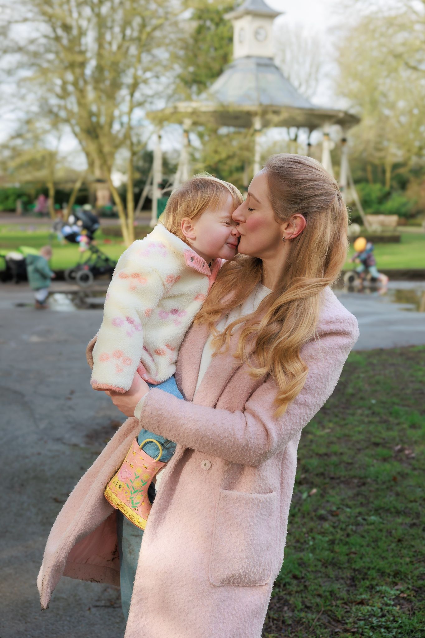 A young woman in a pink coat kisses a toddler she holds in a park with a gazebo backdrop in North Cerney, Cotswolds.