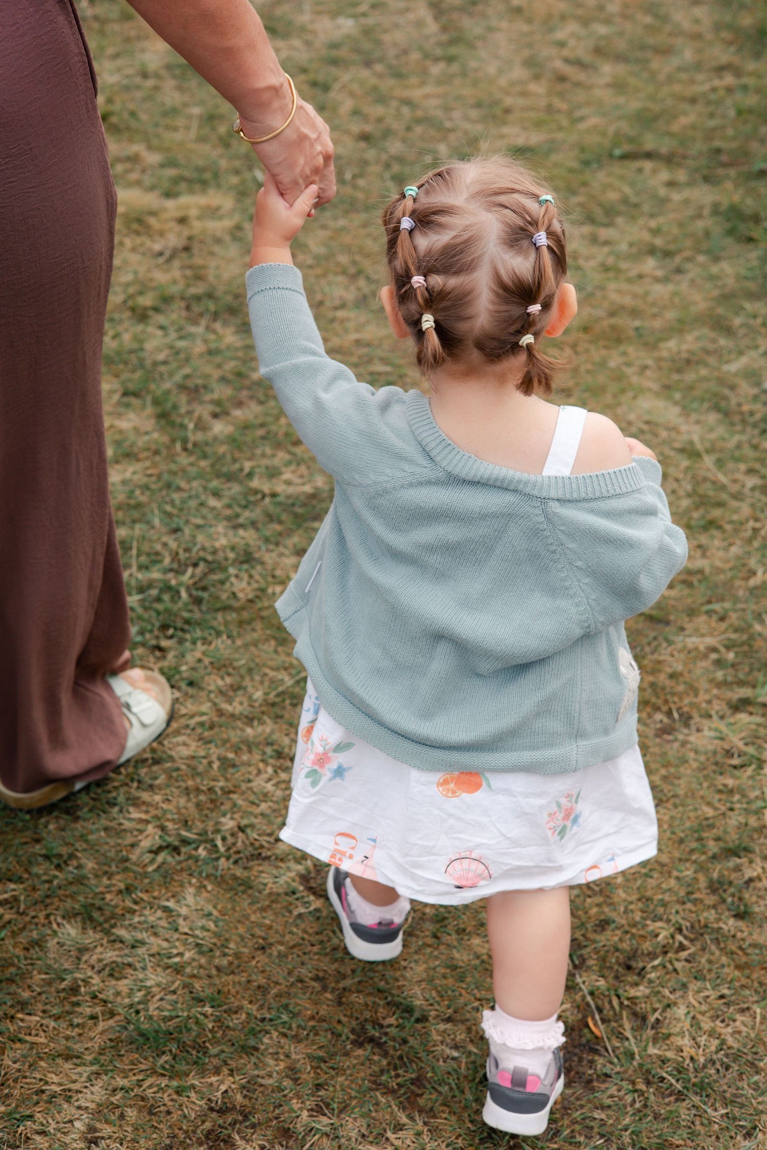 A young girl with braided hair walks with mum, holding her hand in Ascot in a grassy outdoor setting.