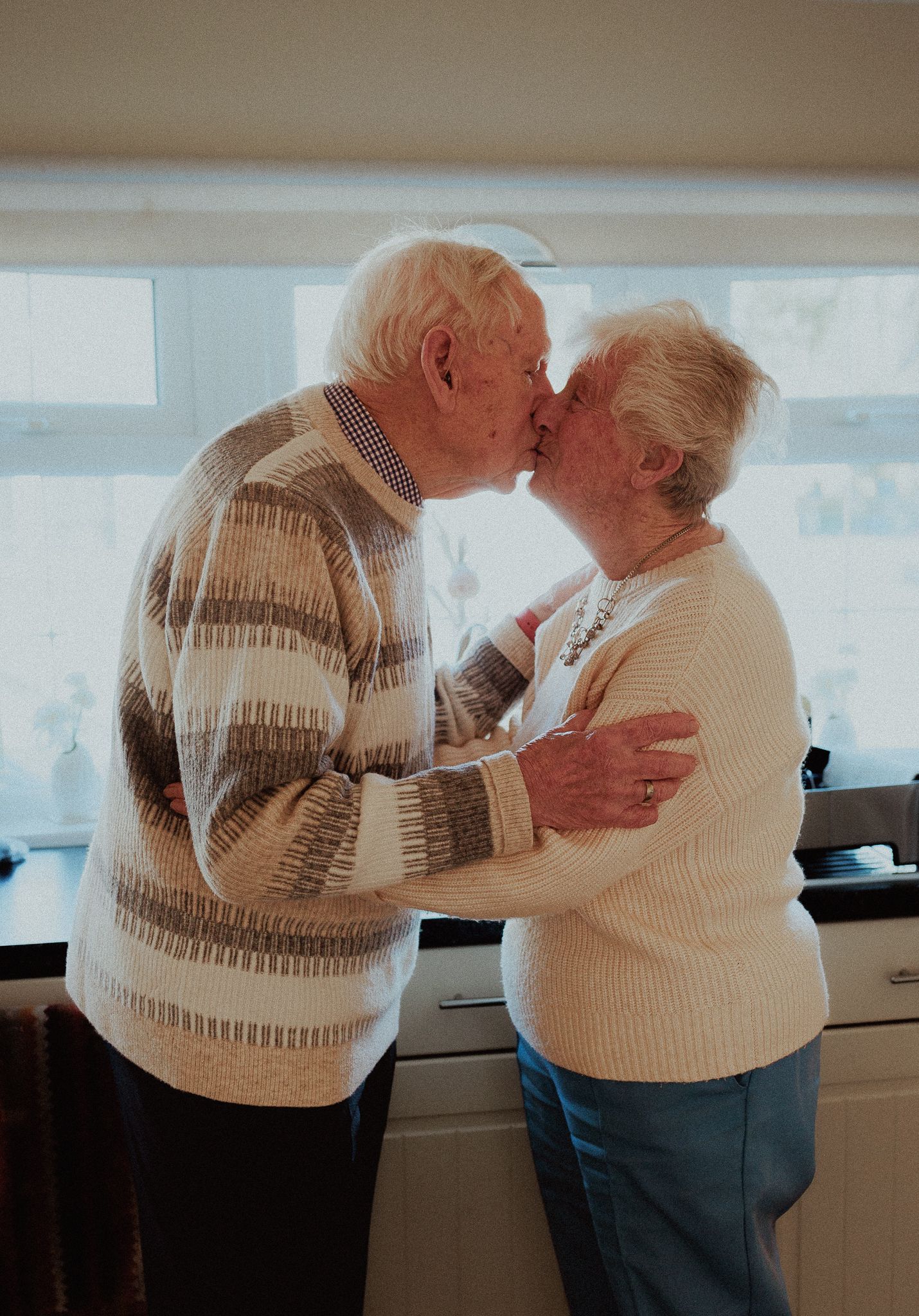 Elderly couple sharing a tender kiss while embracing in a bright kitchen in Castle Combe.