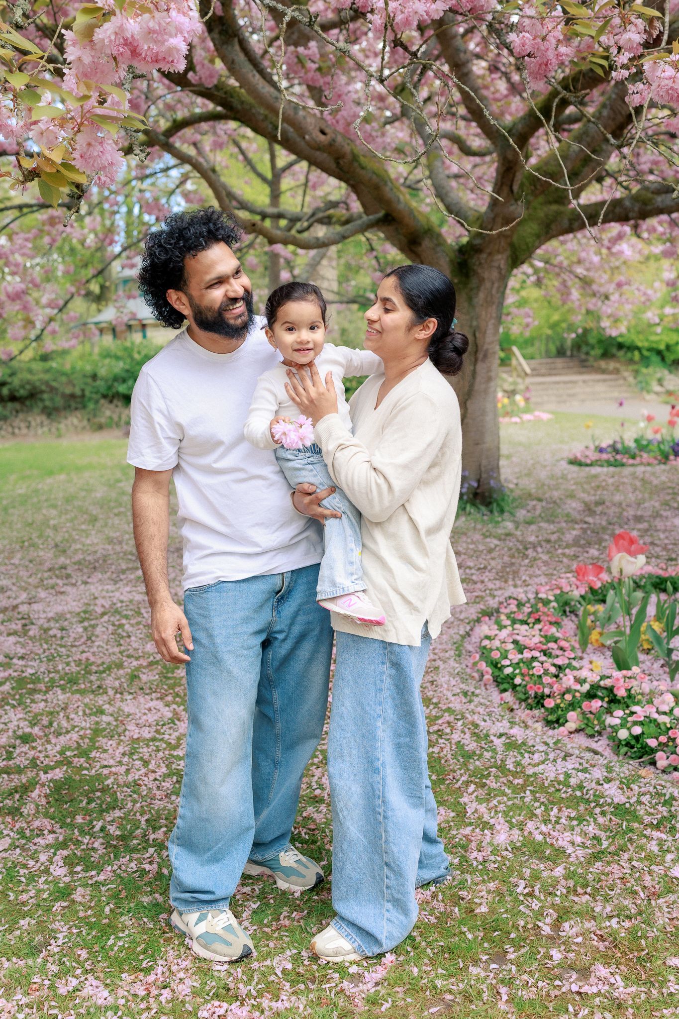 Happy family with toddler posing under pink cherry blossom tree in park during spring, in The Cotswolds.