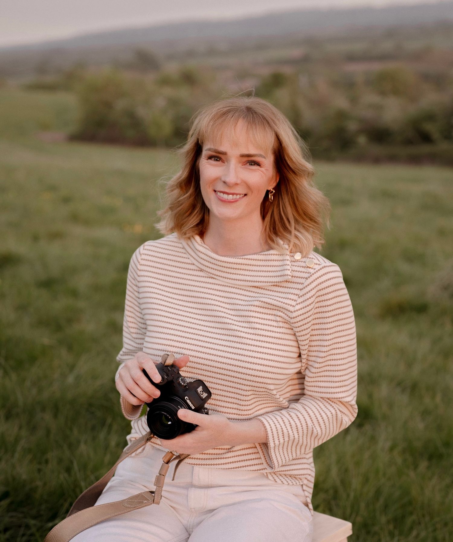 A smiling blonde woman holding a camera sits in a lush green field, wearing a striped beige top in Royal Wootton Bassett.