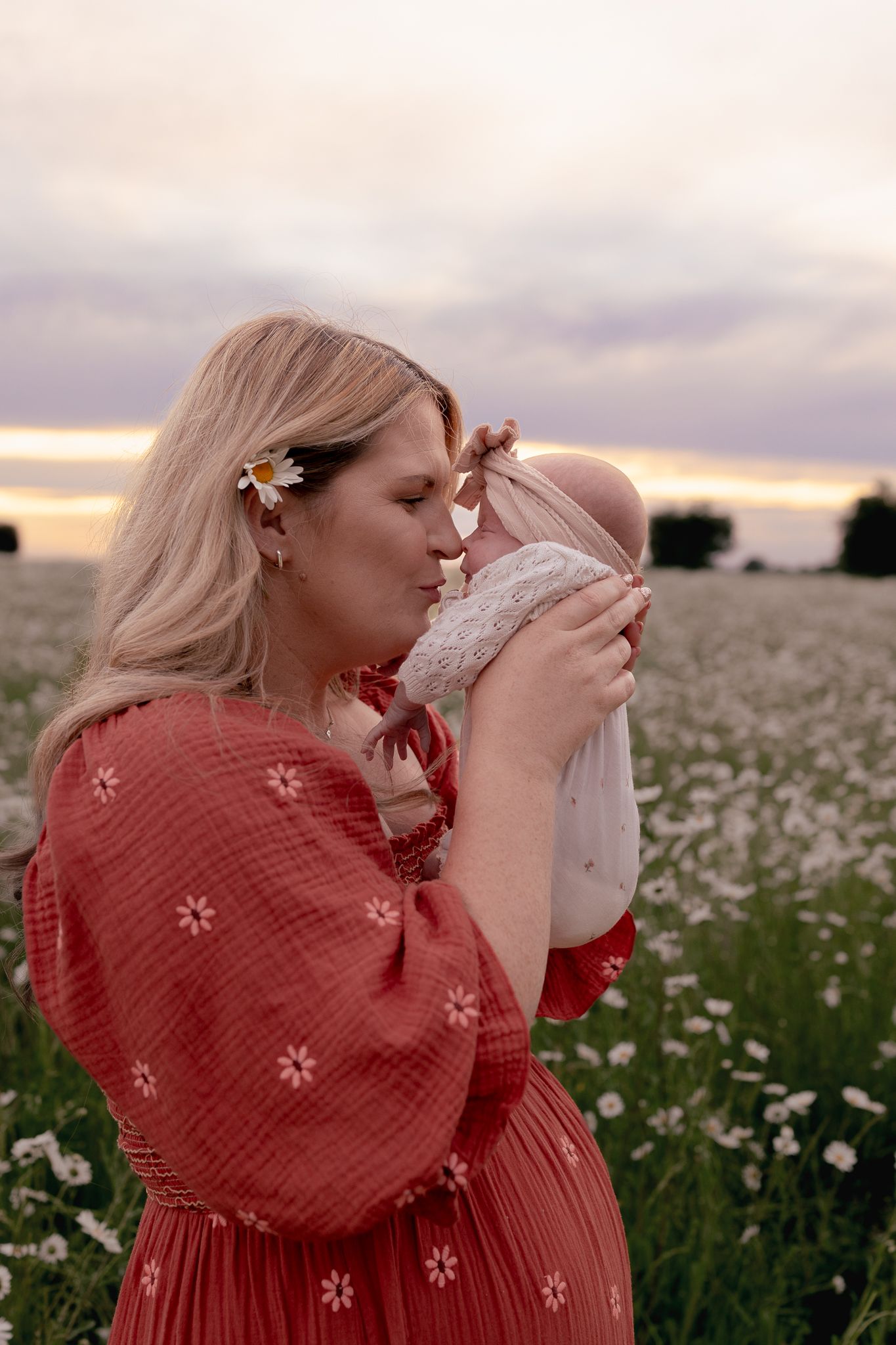A blonde woman in a red floral dress kisses her newborn baby in a blooming daisy field at sunset.