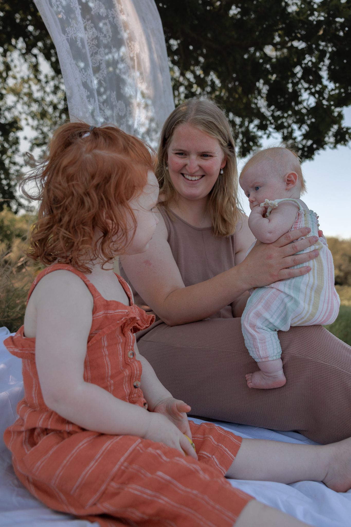 A smiling mother sits outdoors holding her baby while her young daughter in an orange dress sits beside her in Summertown, Oxford.