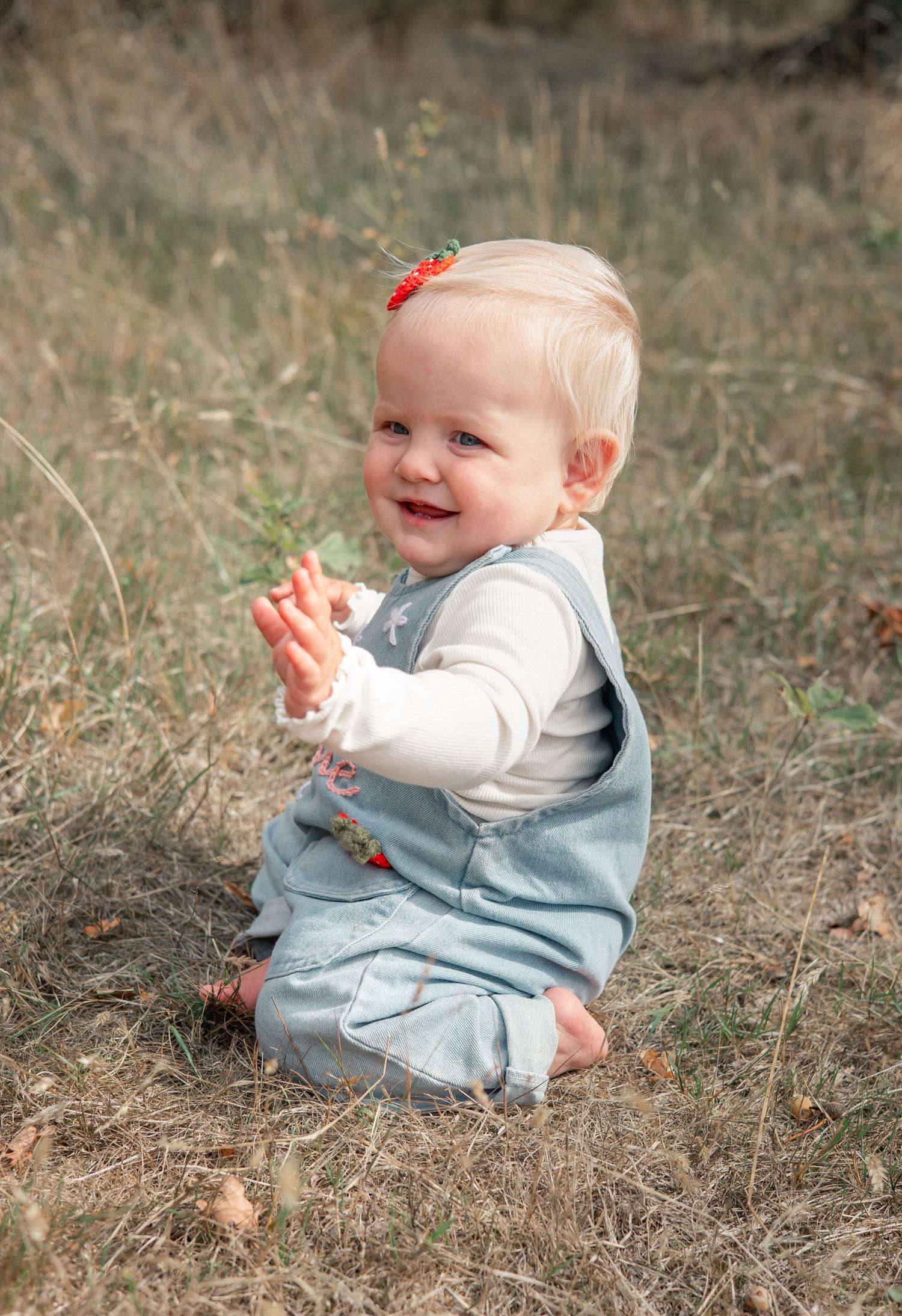 Smiling baby girl in denim overalls and strawberry hair clip clapping hands while sitting on dry grass outdoors.