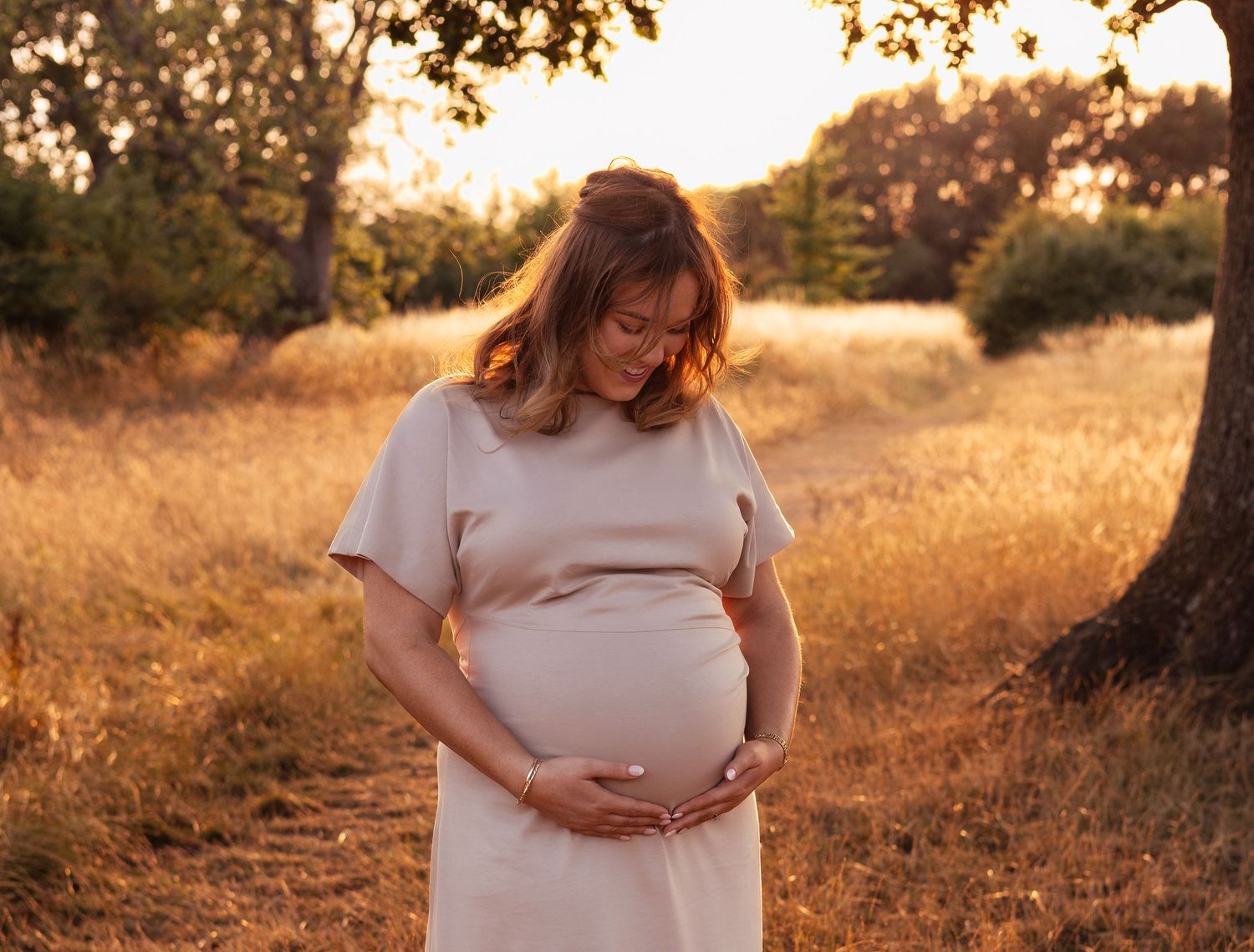 Pregnant woman in beige dress cradling her baby bump in a golden sunset field surrounded by trees in Oxford.