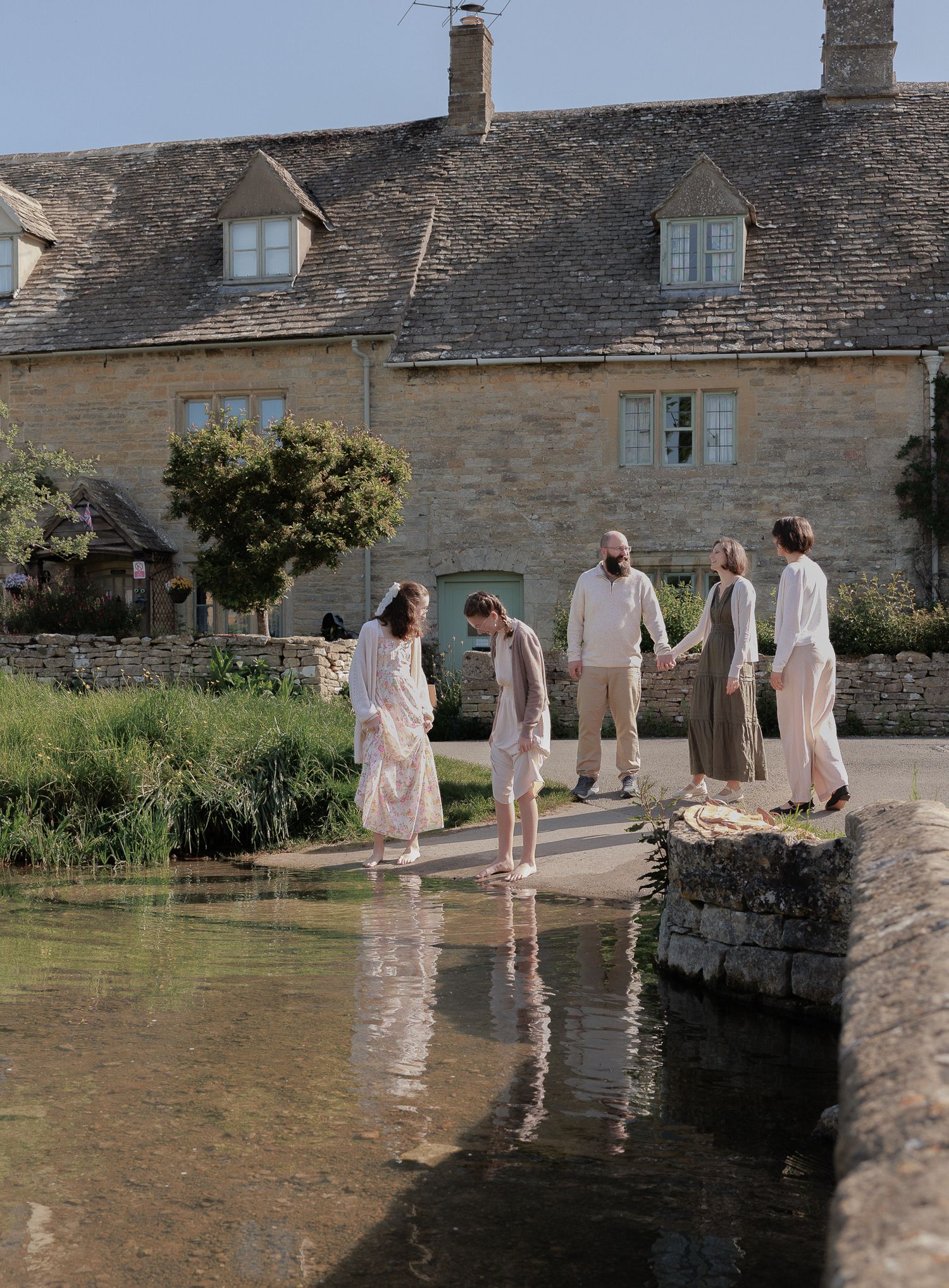 A family wades through a shallow stream beside a charming Cotswolds stone cottage.