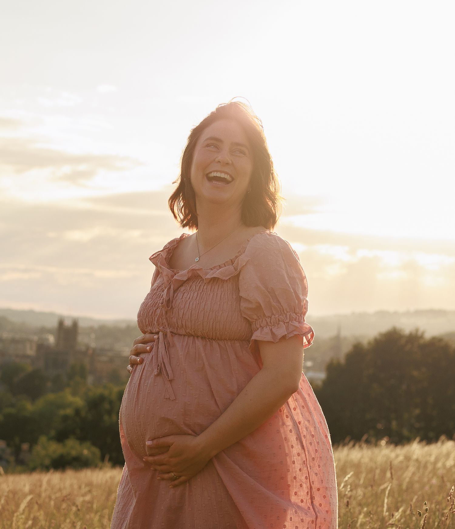 Smiling pregnant woman in pink dress holding baby bump in golden sunset field for a Maternity Photoshoot, Bath, Somerset.