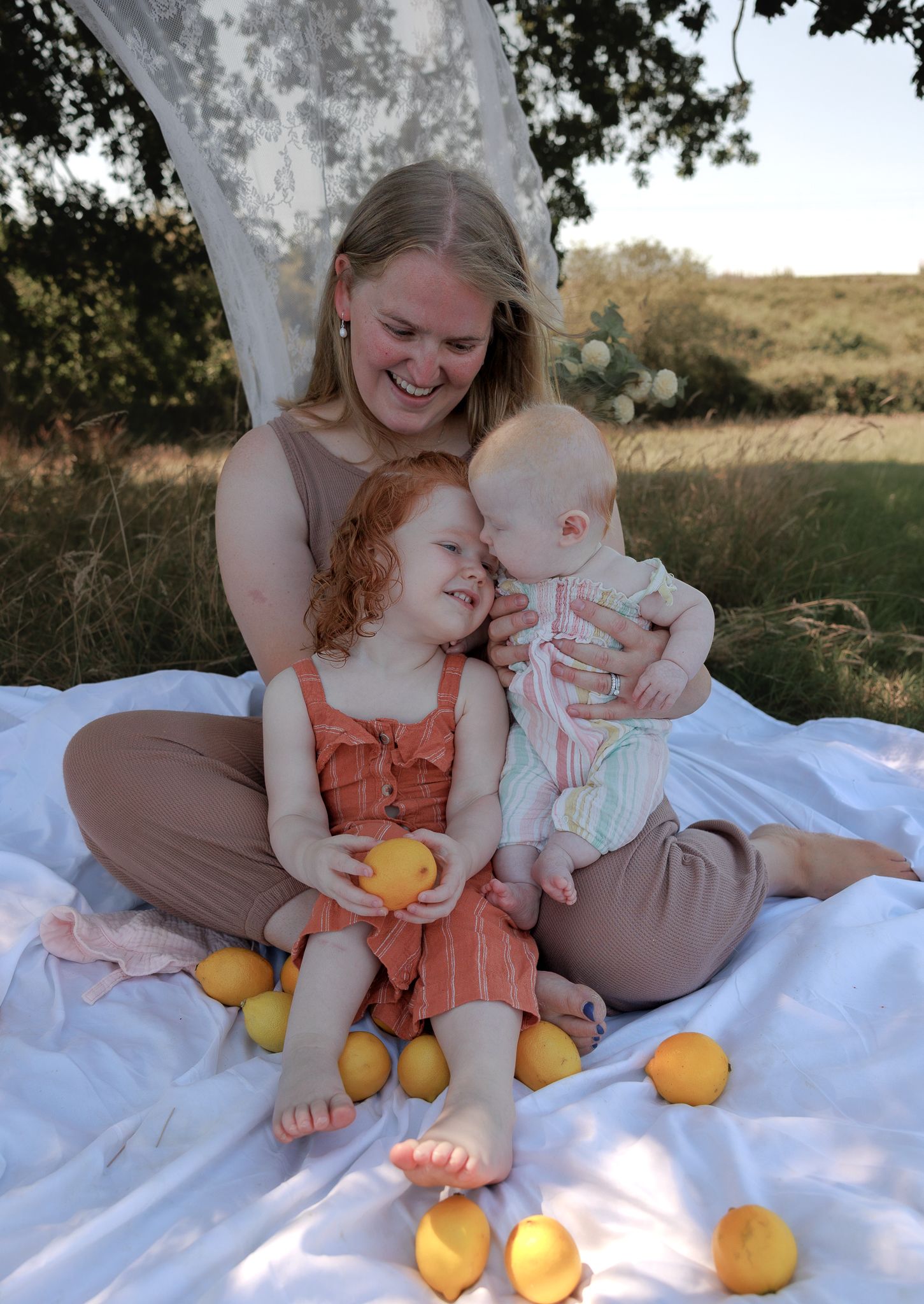 A smiling mother sits on a white blanket outdoors with her two young children surrounded by fresh lemons in Henley-on-Thames, Oxfordshire.