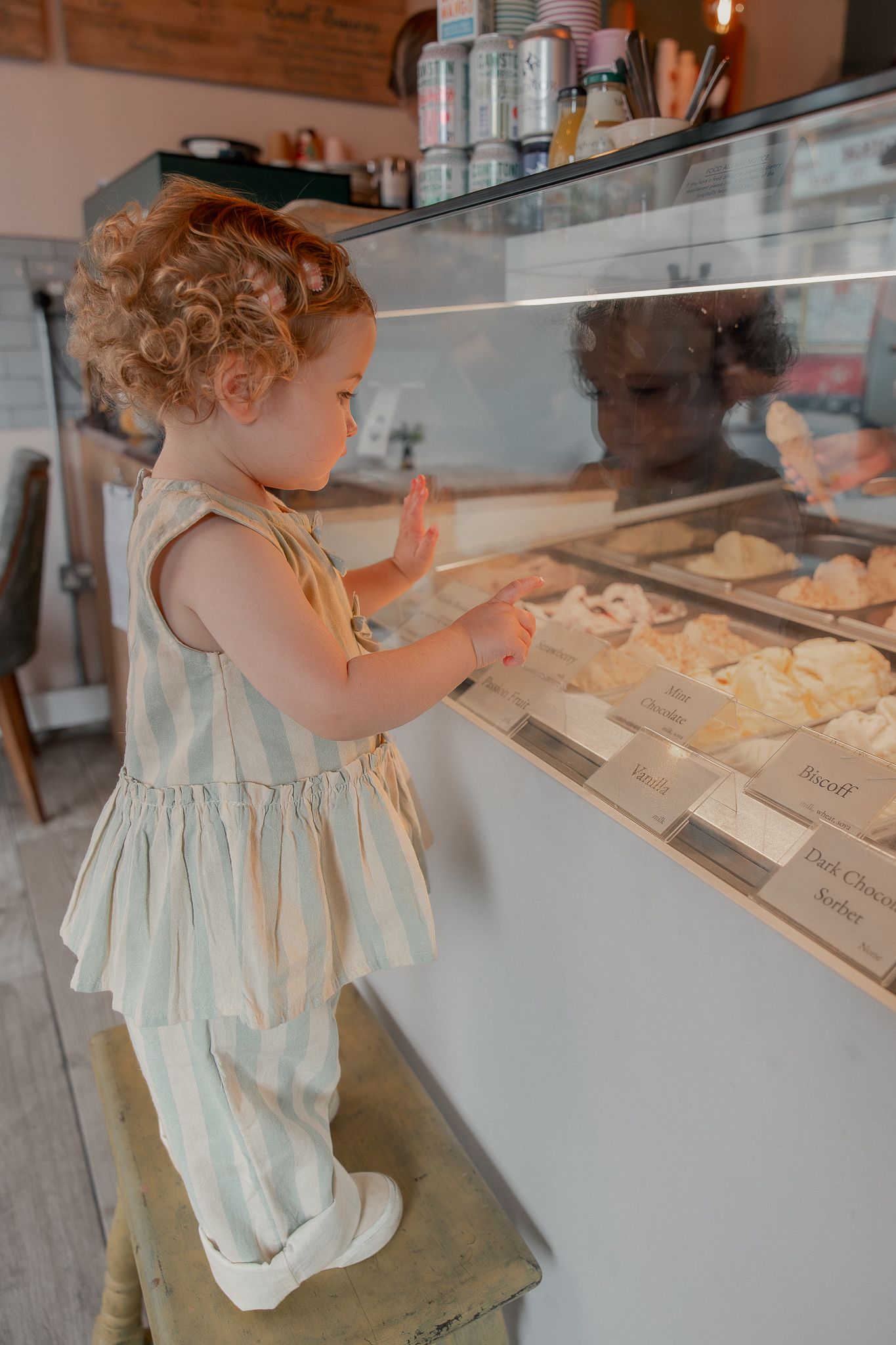 Motherhood photoshoot of curly-haired toddler standing on stool, pointing at pastries in a bakery display case at Cosy Cone Malmesbury.
