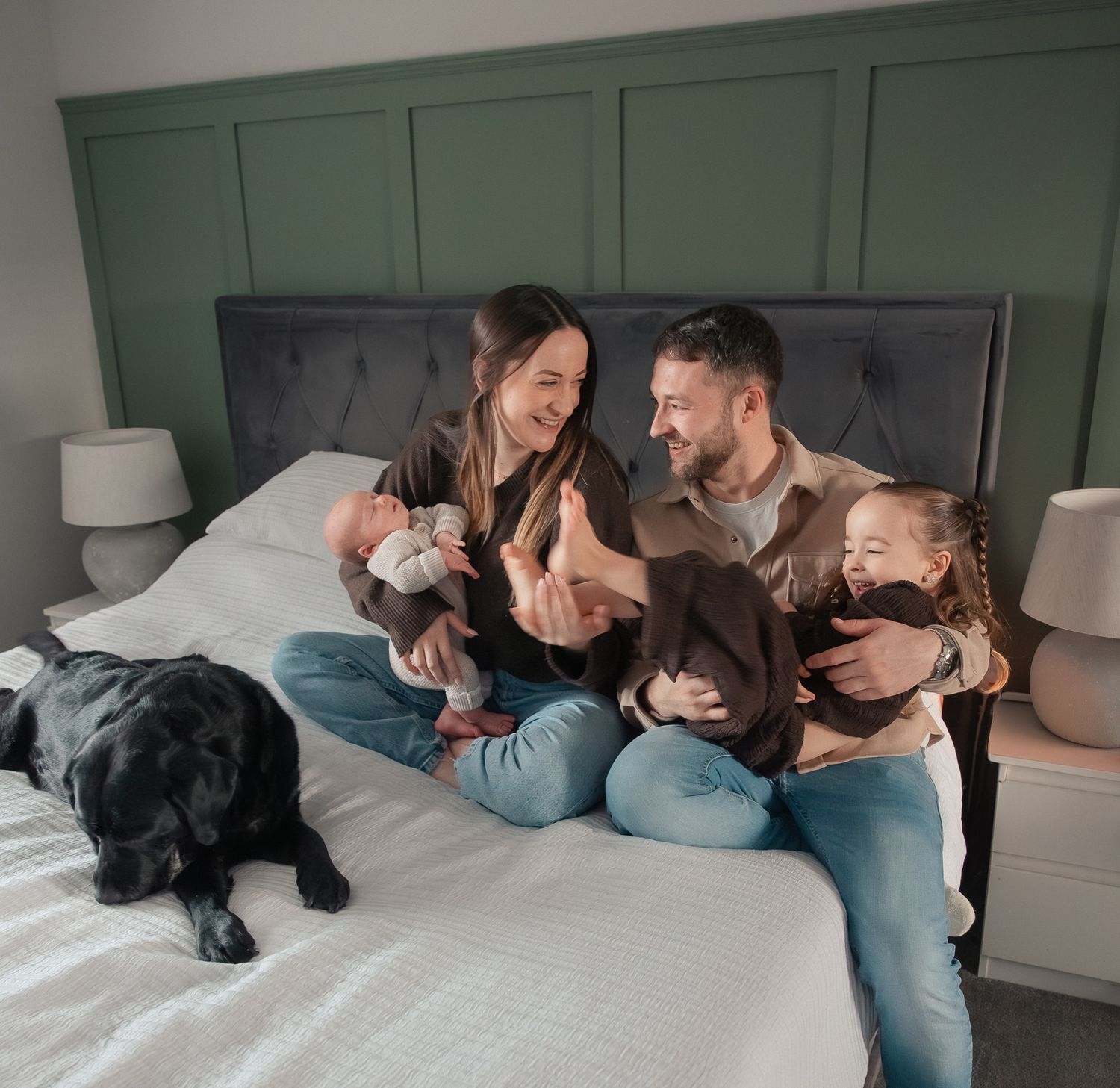 Family with newborn baby and young daughter sitting on bed with black Labrador dog in cosy bedroom, laughing and giggling in Stroud.