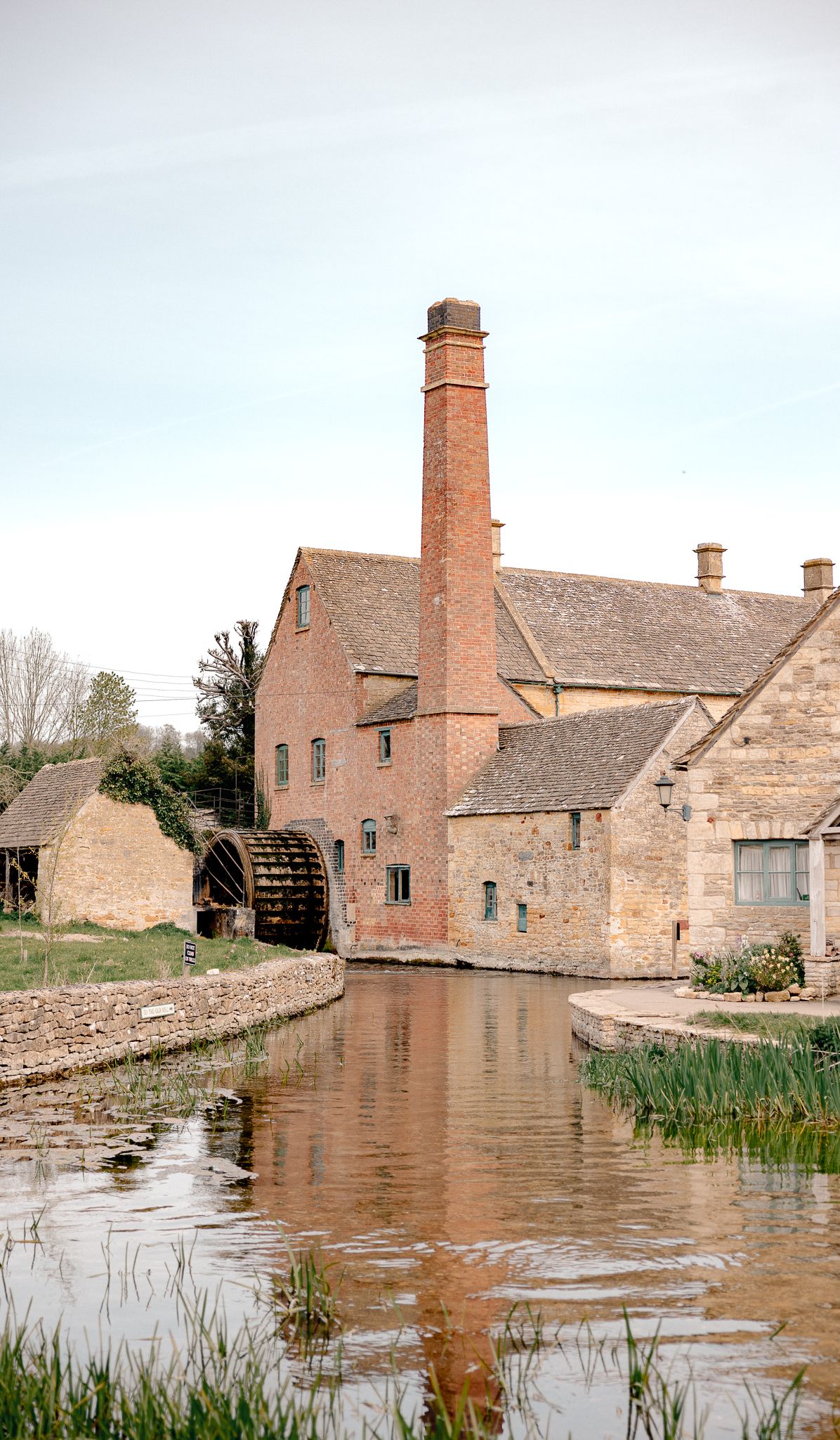 Historic brick watermill with tall chimney reflected in calm millpond, surrounded by stone buildings in rural English countryside, cotswolds
