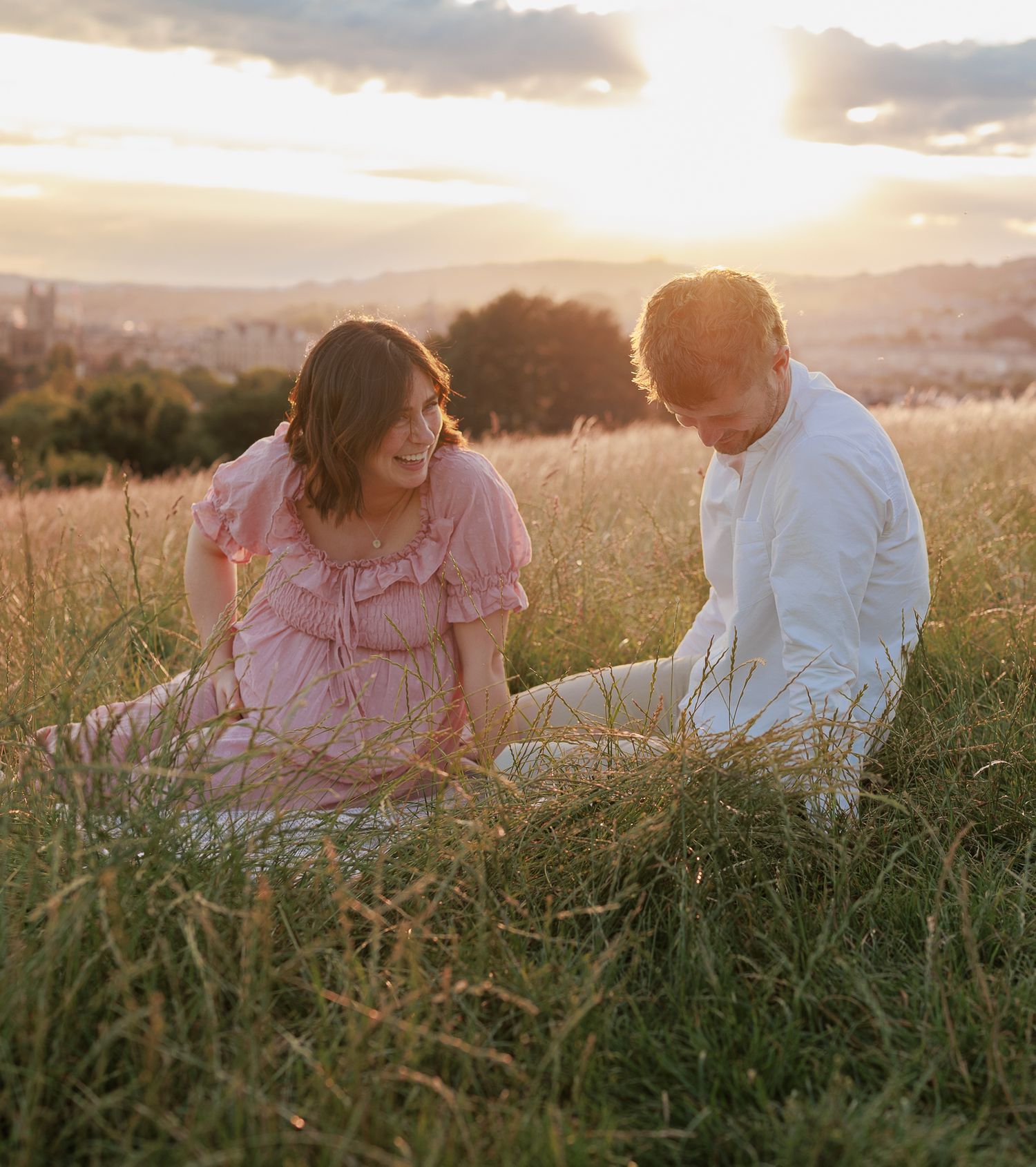 Couple sitting in golden grass field at sunset, woman in pink dress and man in white shirt smiling together in Bristol, UK.