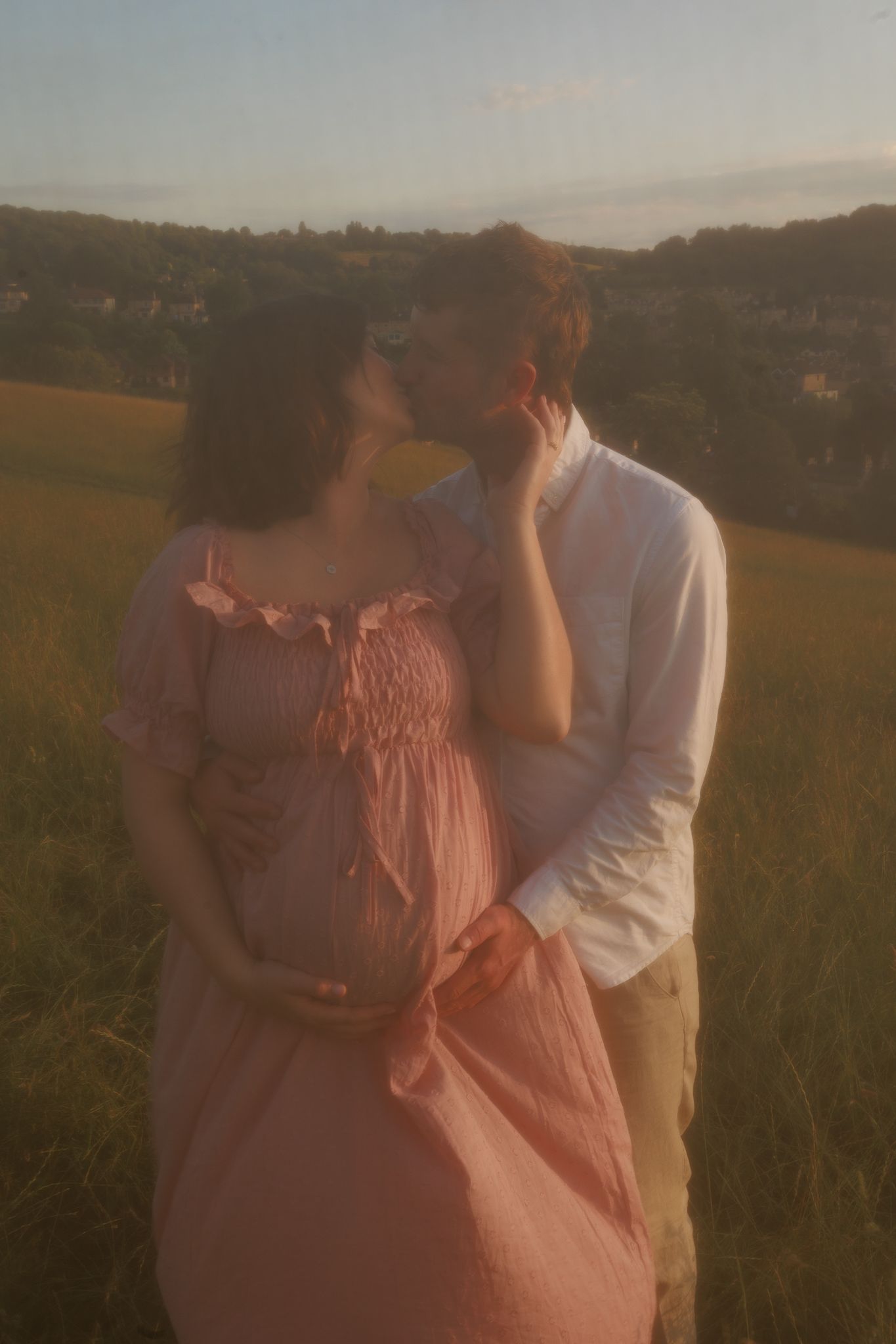 Couple kissing in field at golden hour, pregnant woman in pink dress held tenderly for a Vintage photoshoot in Bath, England.