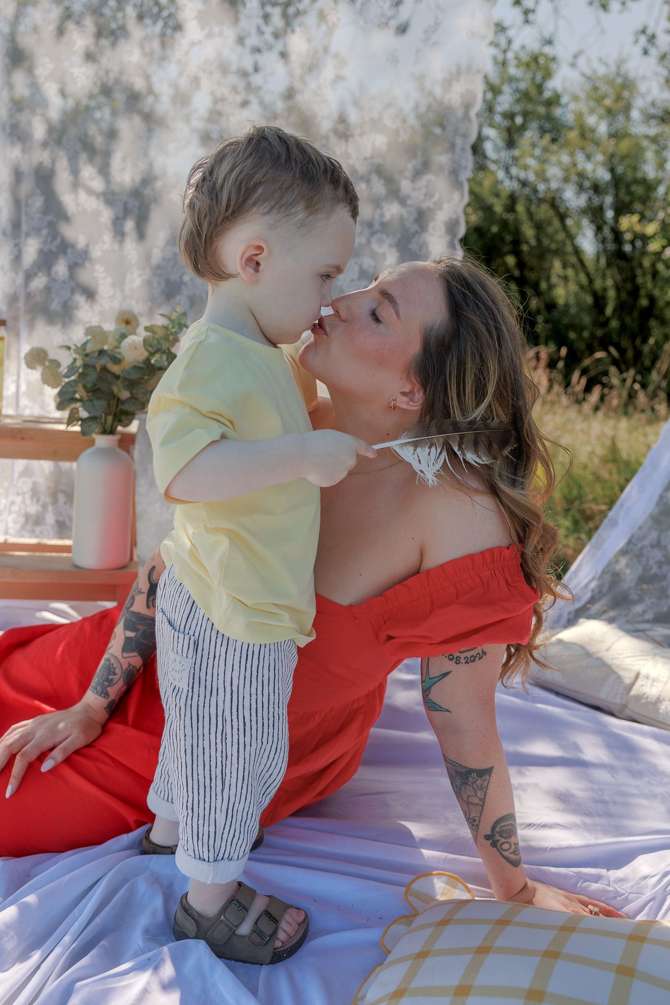 A mother in a red dress shares a tender kiss with her toddler son in a sunny outdoor setting in Fairford, Cotswolds.
