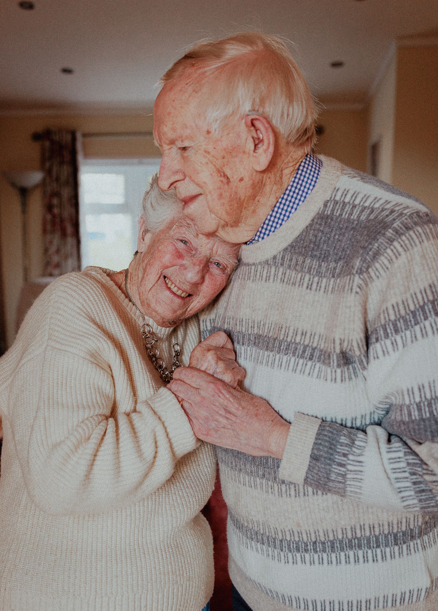 Elderly couple dancing together indoors, smiling warmly while holding hands in matching cream sweaters in Bourton-on-the-water.
