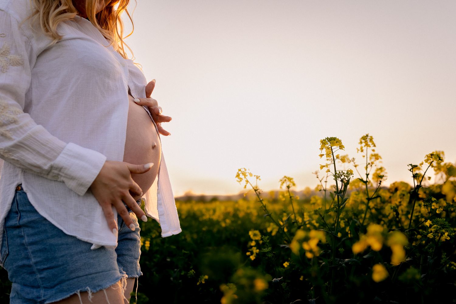 Pregnant woman in white shirt and denim shorts cradling baby bump in golden sunlit field of yellow flowers in Wallingford.