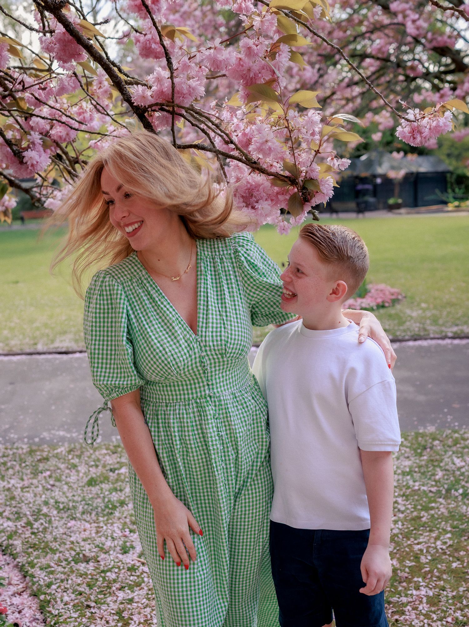 Woman in green gingham dress and boy in white shirt smiling under pink cherry blossom tree in park in Clevedon.