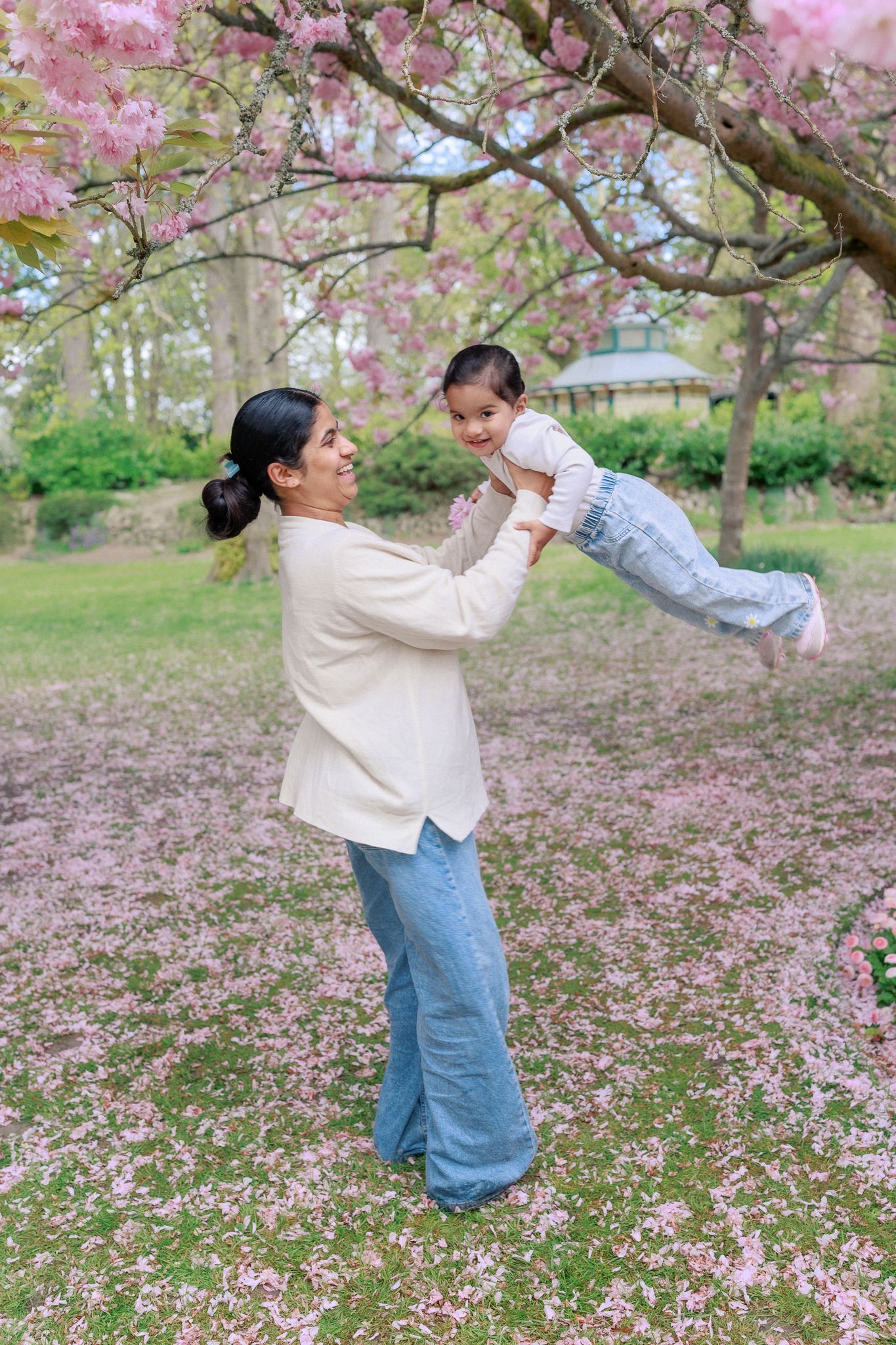A mother joyfully lifts her toddler under blooming pink cherry blossom trees in a lush green park in Bath, Somerset.