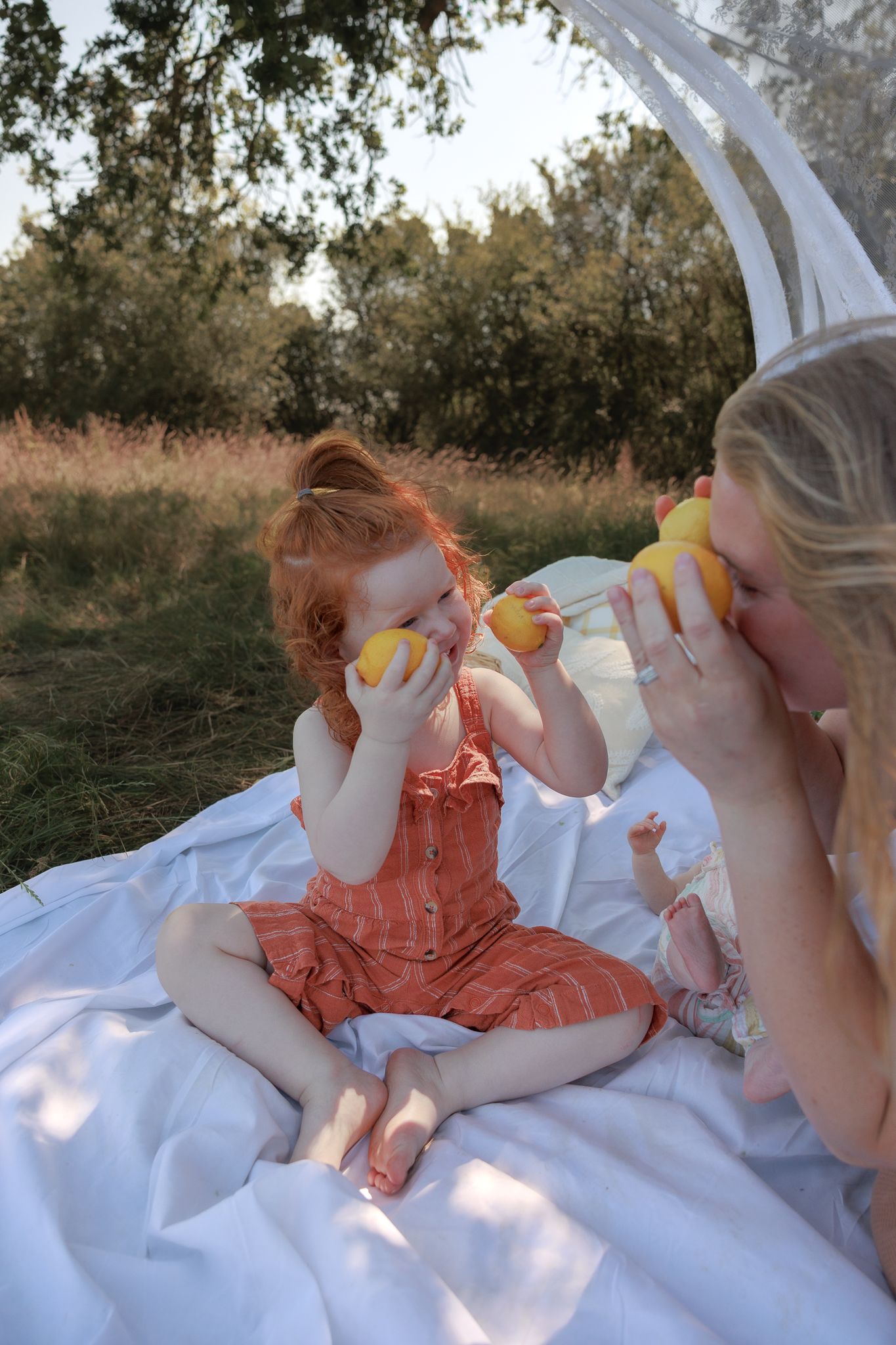 Redhead toddler girl and blonde mother smelling lemons together on white blanket during outdoor picnic in Henley-on-Thames, Oxfordshire.