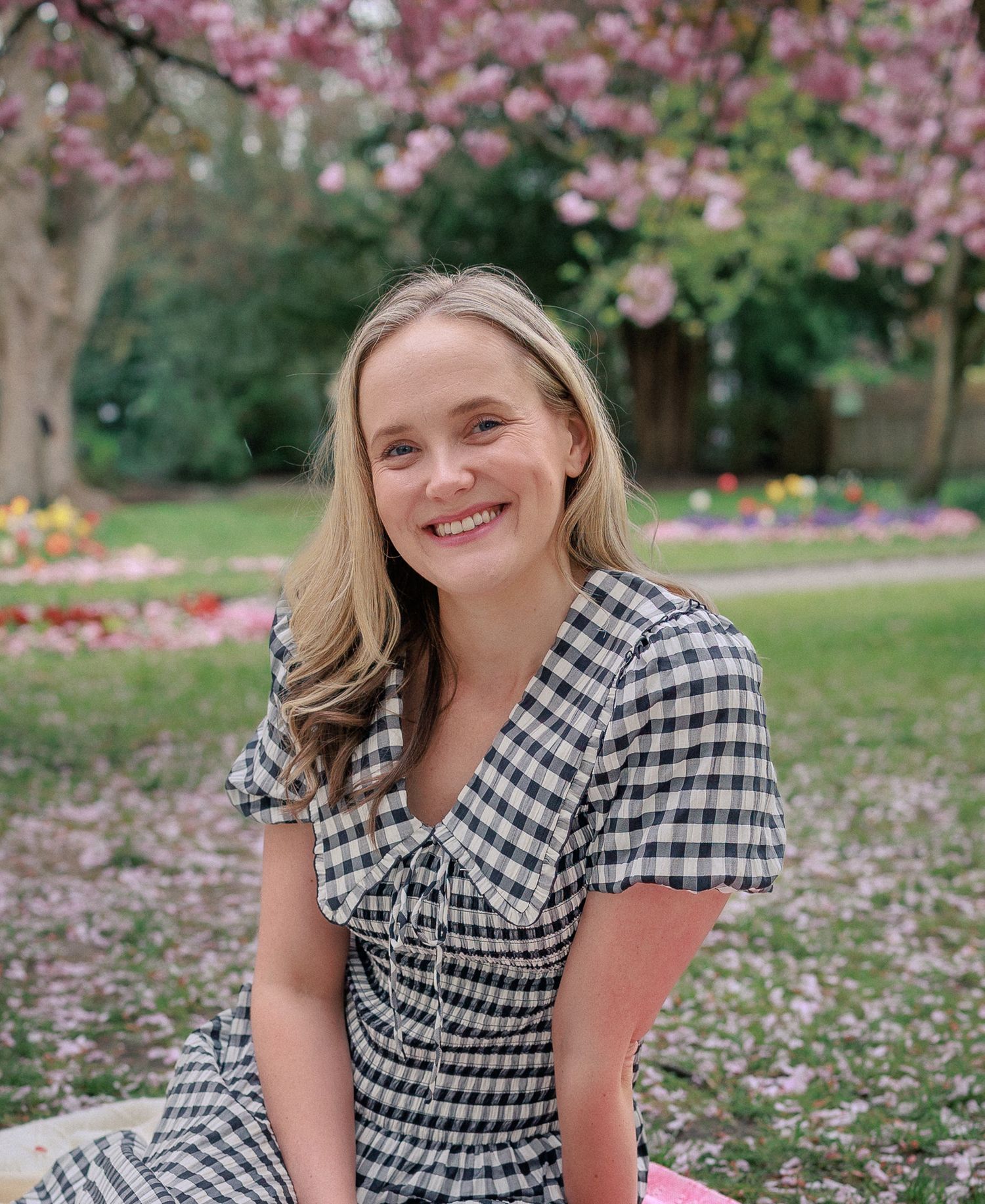 Smiling blonde woman in gingham dress sitting outdoors under blooming cherry blossom tree in colourful garden park in Westminster.