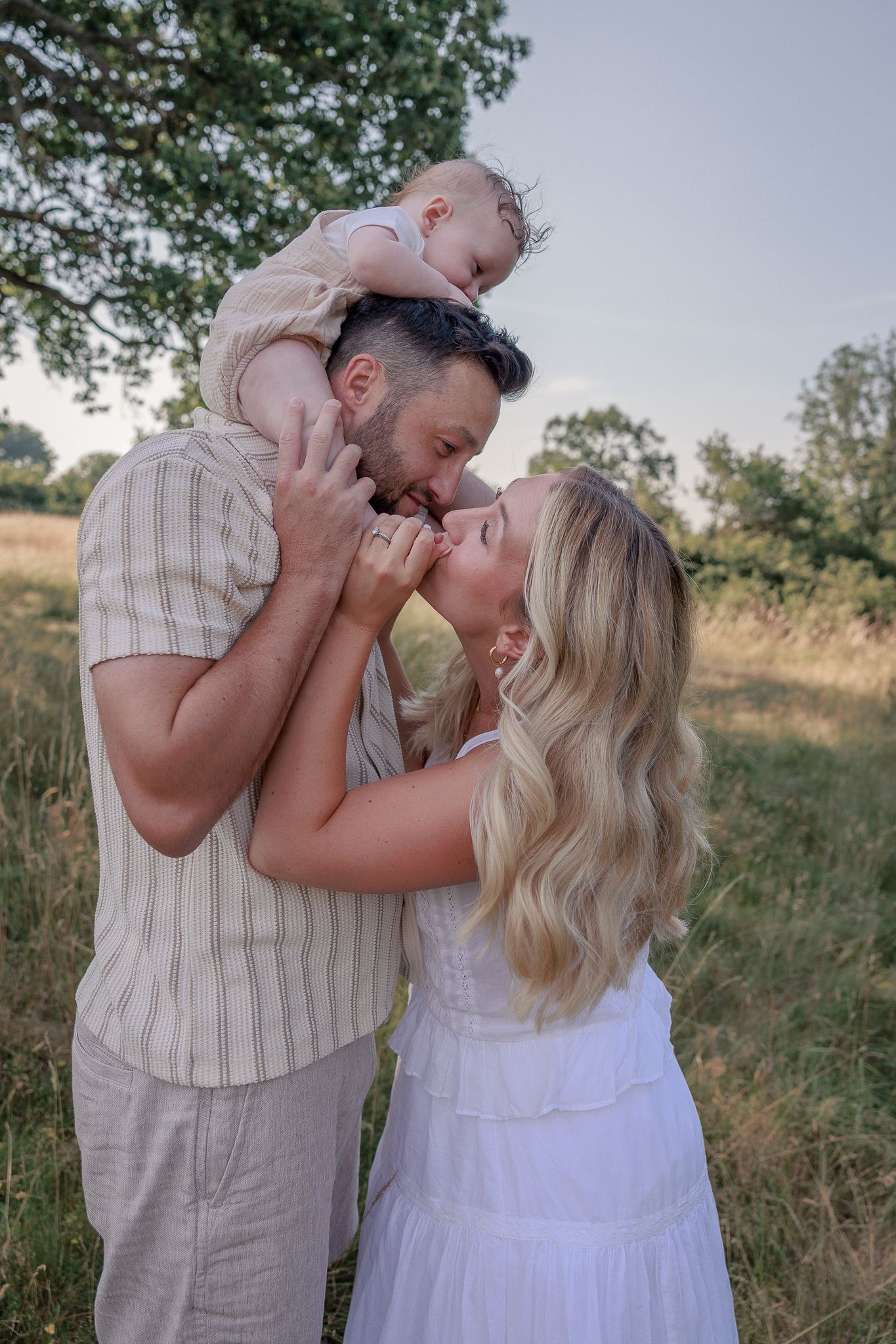 Happy couple kissing while man holds baby overhead in a sunny outdoor field setting in South Cerney, Cotswolds.