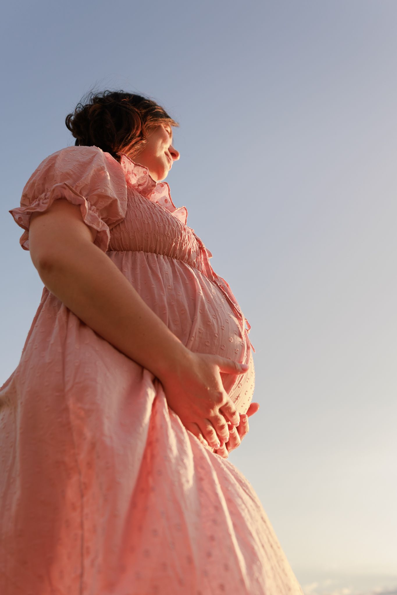 Pregnant woman in pink dress cradling her baby bump outdoors against a soft blue sky at sunset during Maternity Photoshoot in Reading.