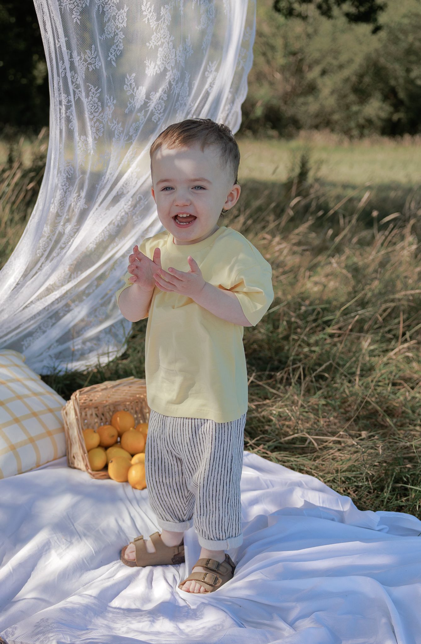 A happy toddler in yellow shirt and striped pants claps hands near lemons on a white blanket outdoors in Fairford, Cotswolds.