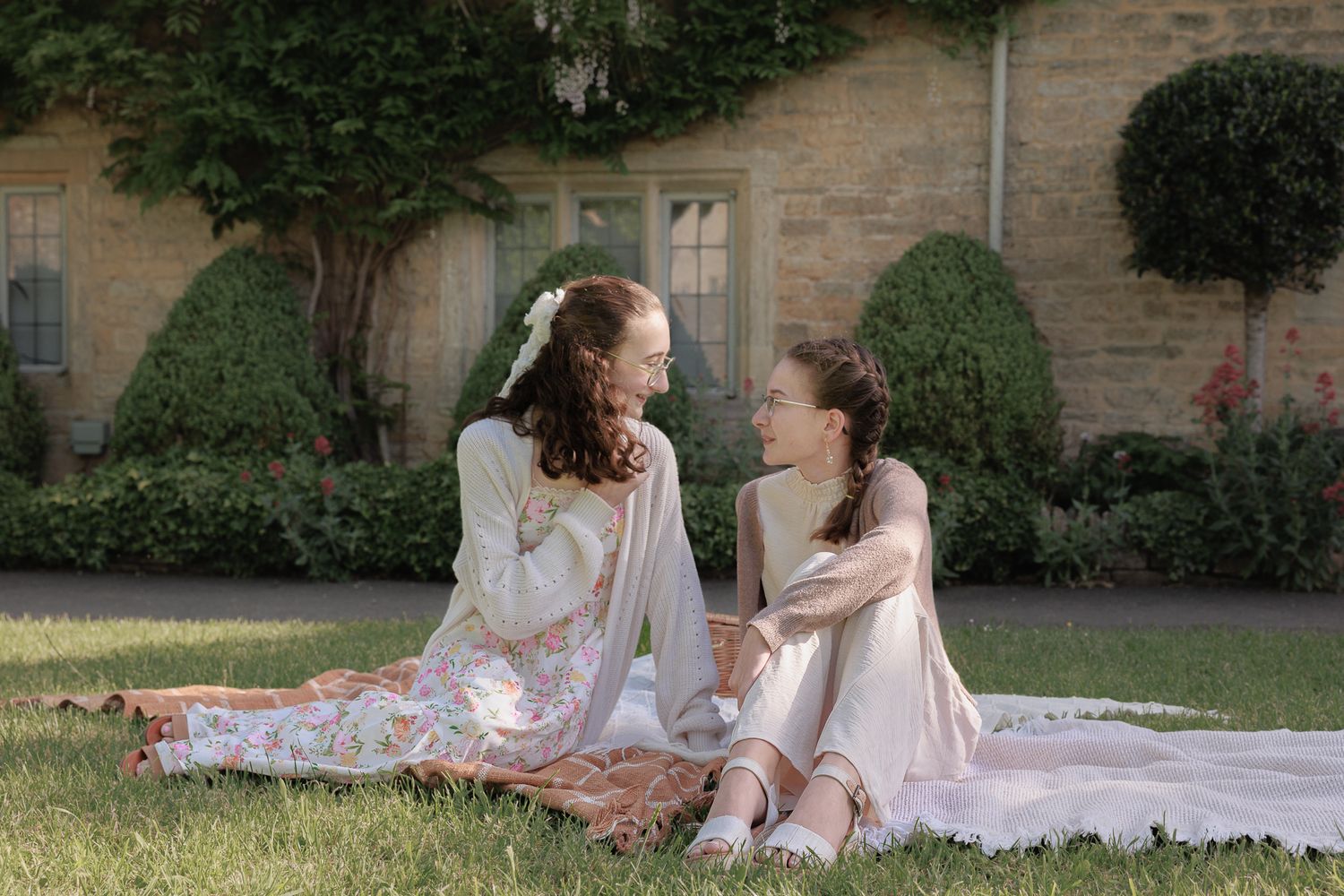 Two young women in floral dresses sit on a blanket in a lush garden, sharing a joyful conversation.
