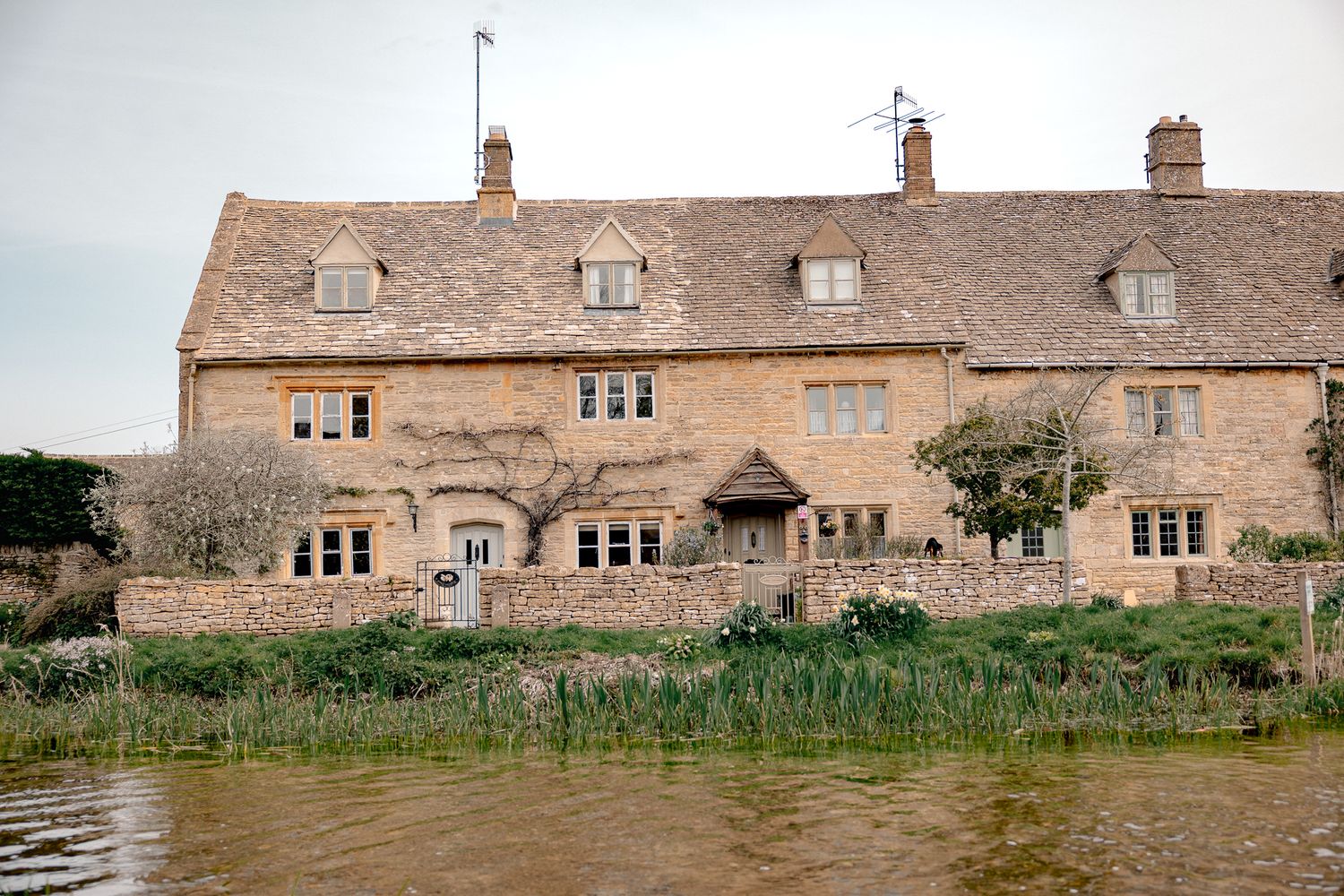 Historic Cotswold stone cottage beside a tranquil river, featuring climbing plants and traditional English architecture in Lower Slaughter.