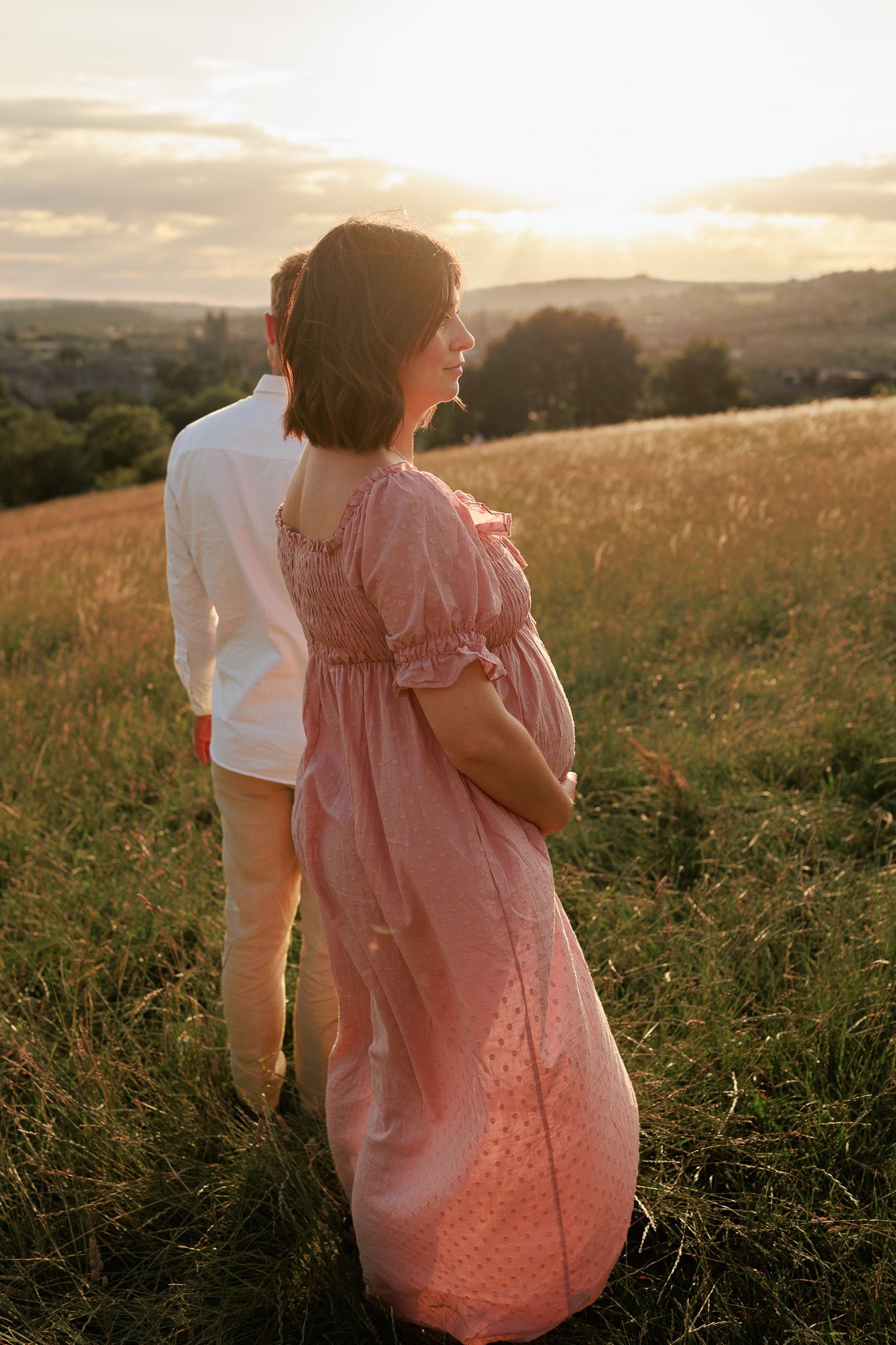 Pregnant woman during Maternity Photoshoot in pink dress stands with partner viewing golden sunset over rolling fields over Bath, Somerset.