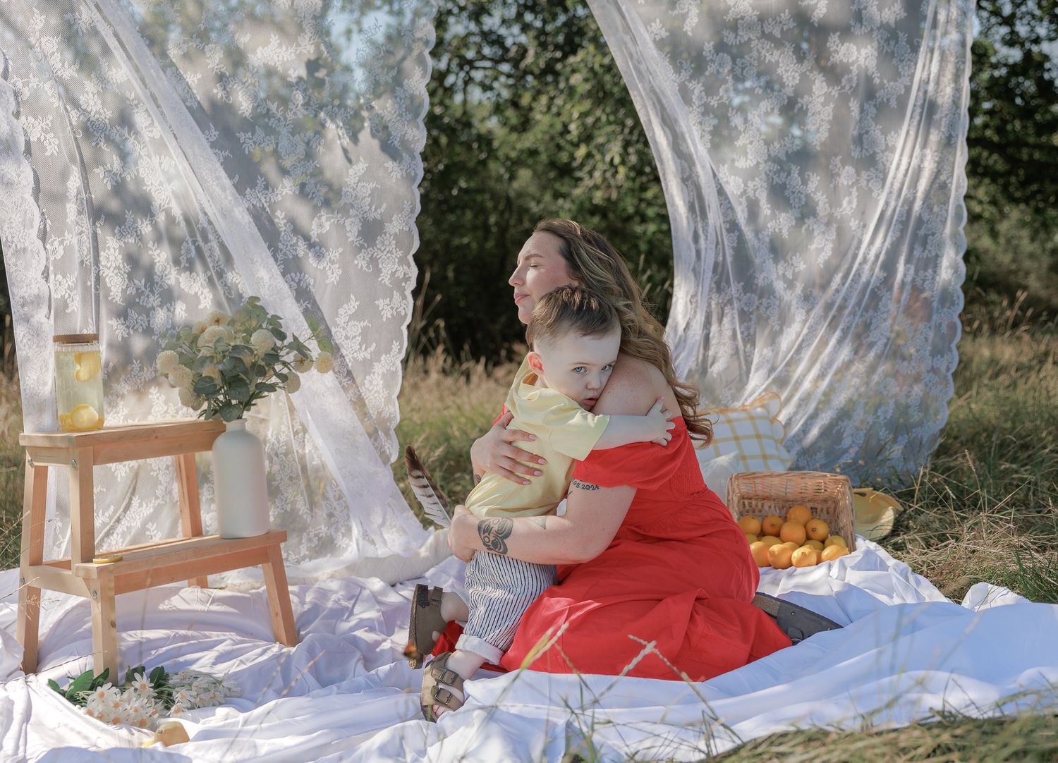 A woman in a red dress hugs a child on a white blanket during an outdoor picnic with sheer curtains in Fairford, Cotswolds.