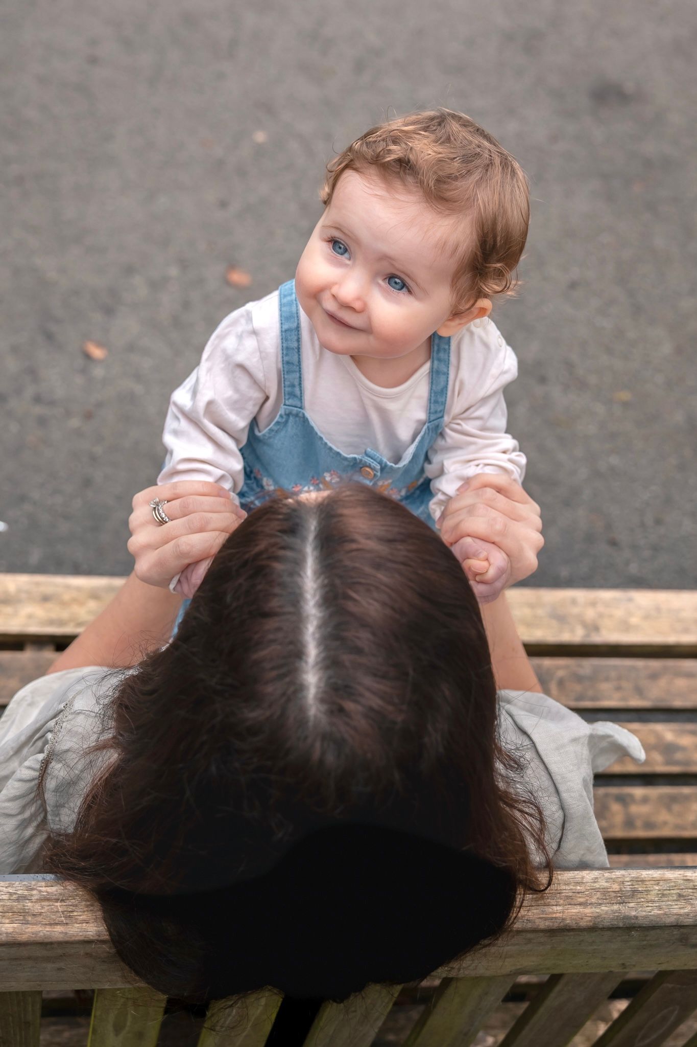 A smiling baby in denim overalls sits on a bench with a dark-haired woman viewed from above in a park in South Cerney, Cotswolds.