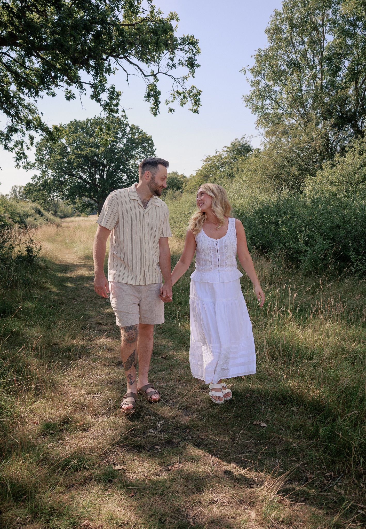Couple holding hands walking along a sunny countryside path surrounded by lush green trees and tall grass in Stow, Cotswolds.