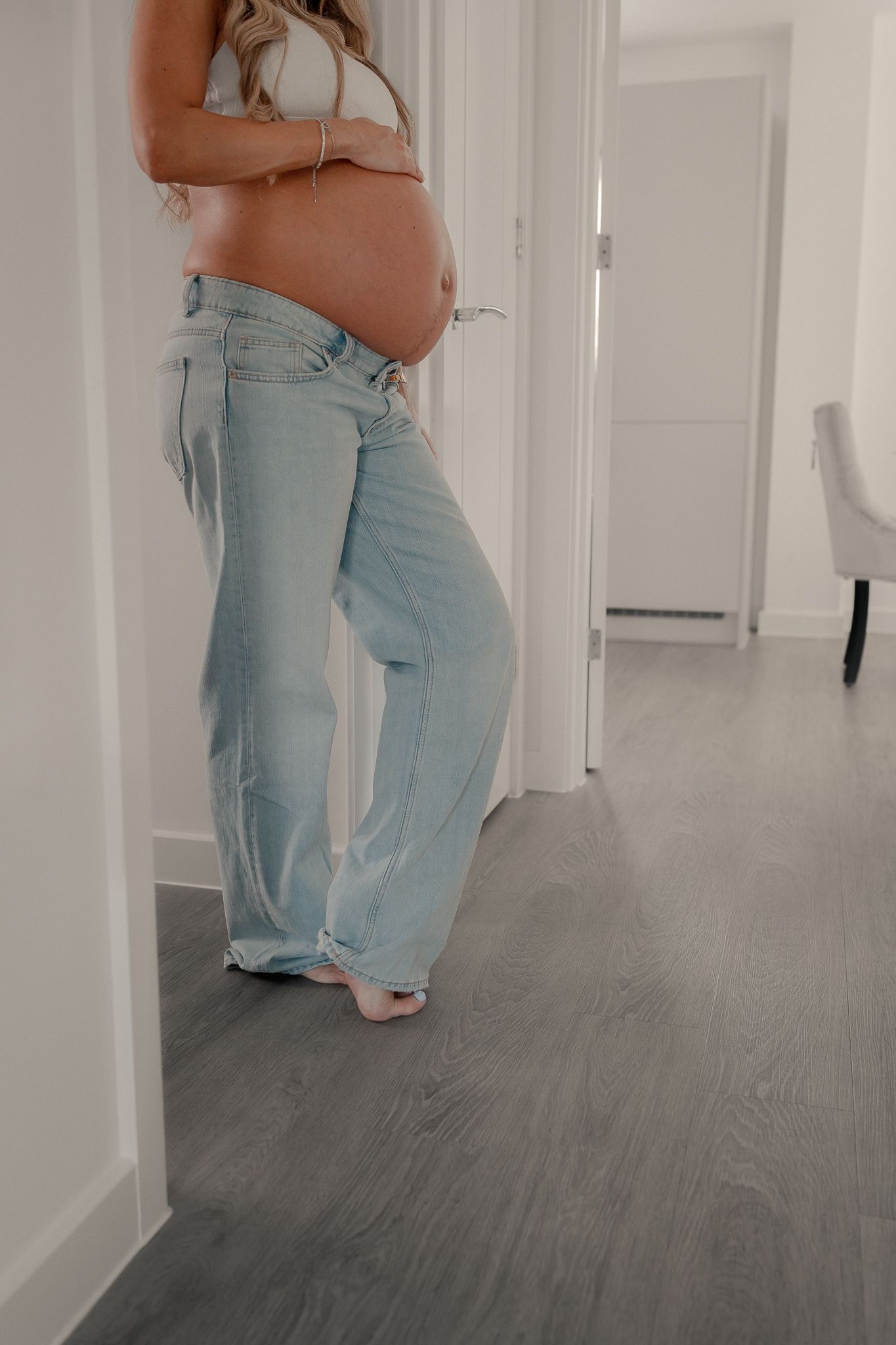 Pregnant woman showing baby bump in light wash wide-leg jeans and white crop top, standing in a bright hallway in Ramsbury.