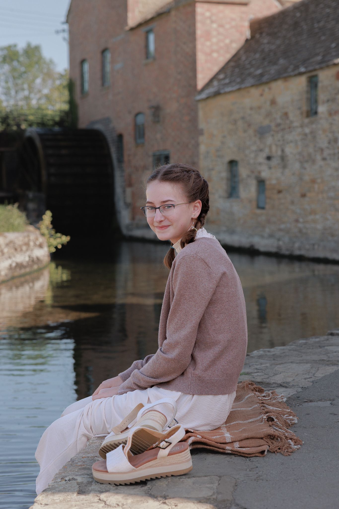 Young woman with glasses sits by a canal near 'The Old Mill' in Lower Slaughter.