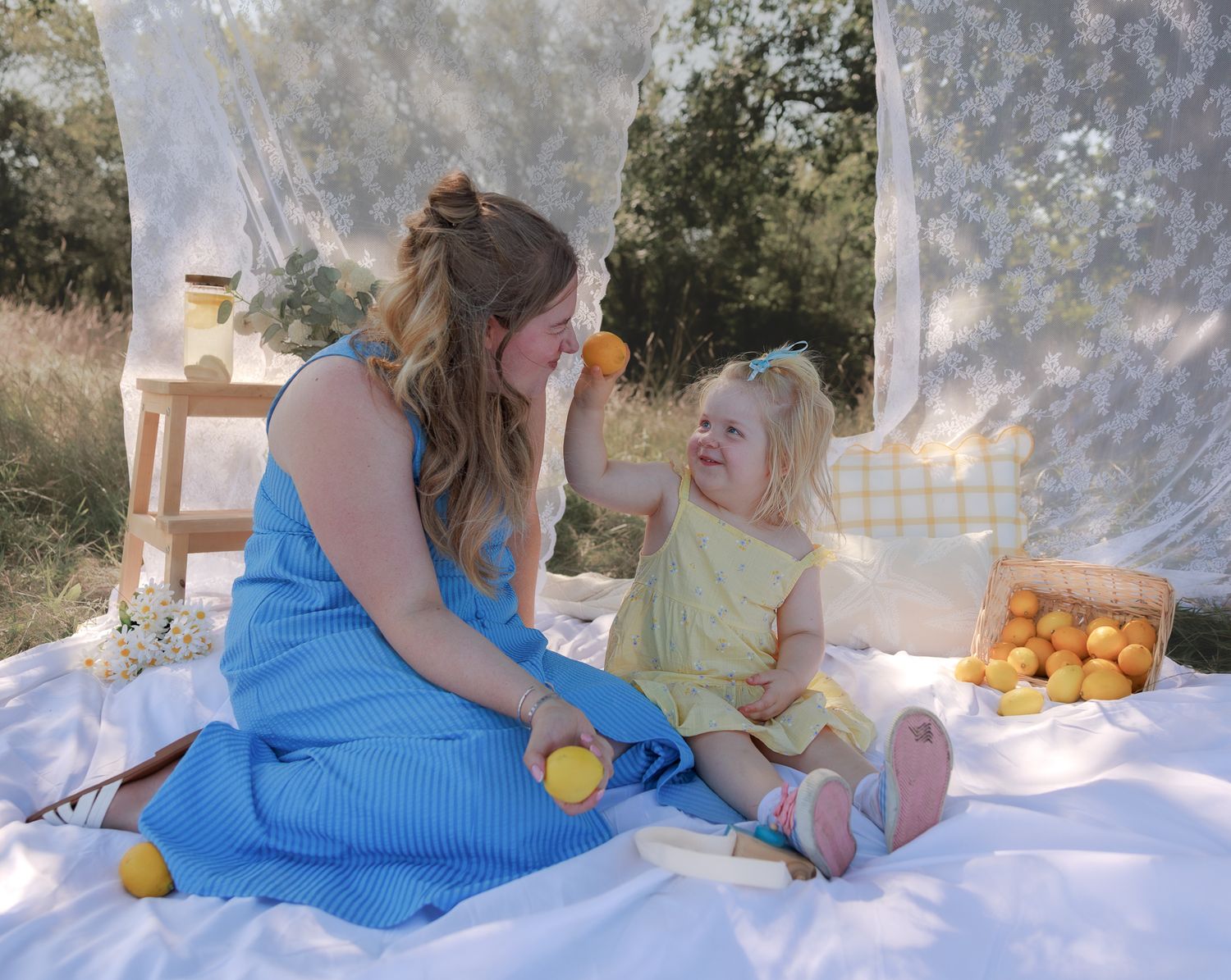 A mother and toddler girl enjoy an outdoor picnic with oranges on a white blanket surrounded by sheer white curtains in Bibury, Cotswolds.