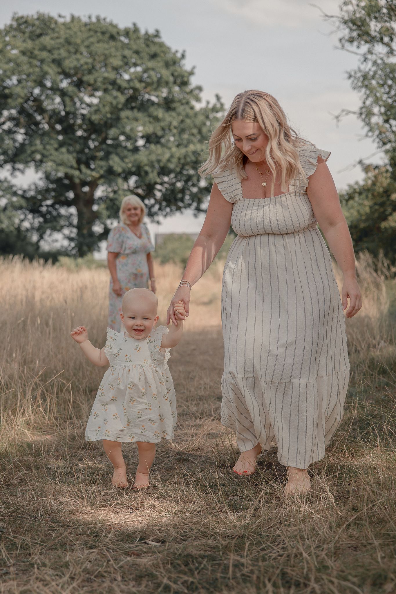 A mother in a striped dress holds her baby girl's hand while walking barefoot through a golden field.