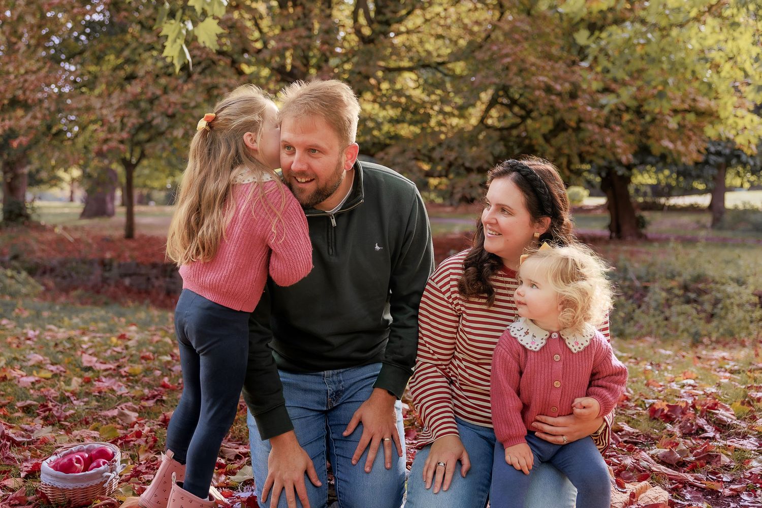 Family of four enjoying autumn outdoors, parents smiling as daughters share a sweet moment among colorful fallen leaves.
