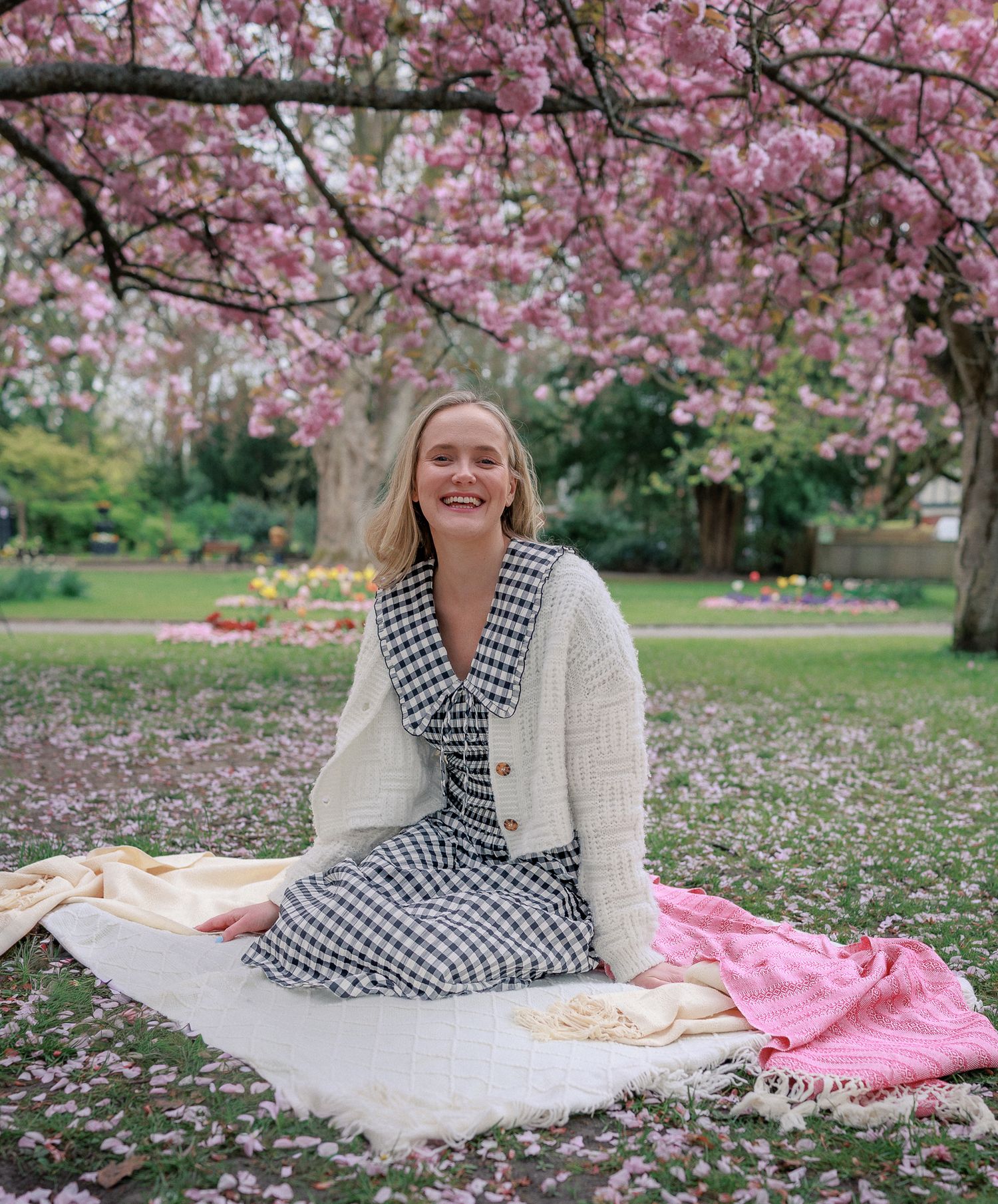 Woman in white cardigan and gingham dress sitting on blanket under pink cherry blossom tree in park in Northleach.