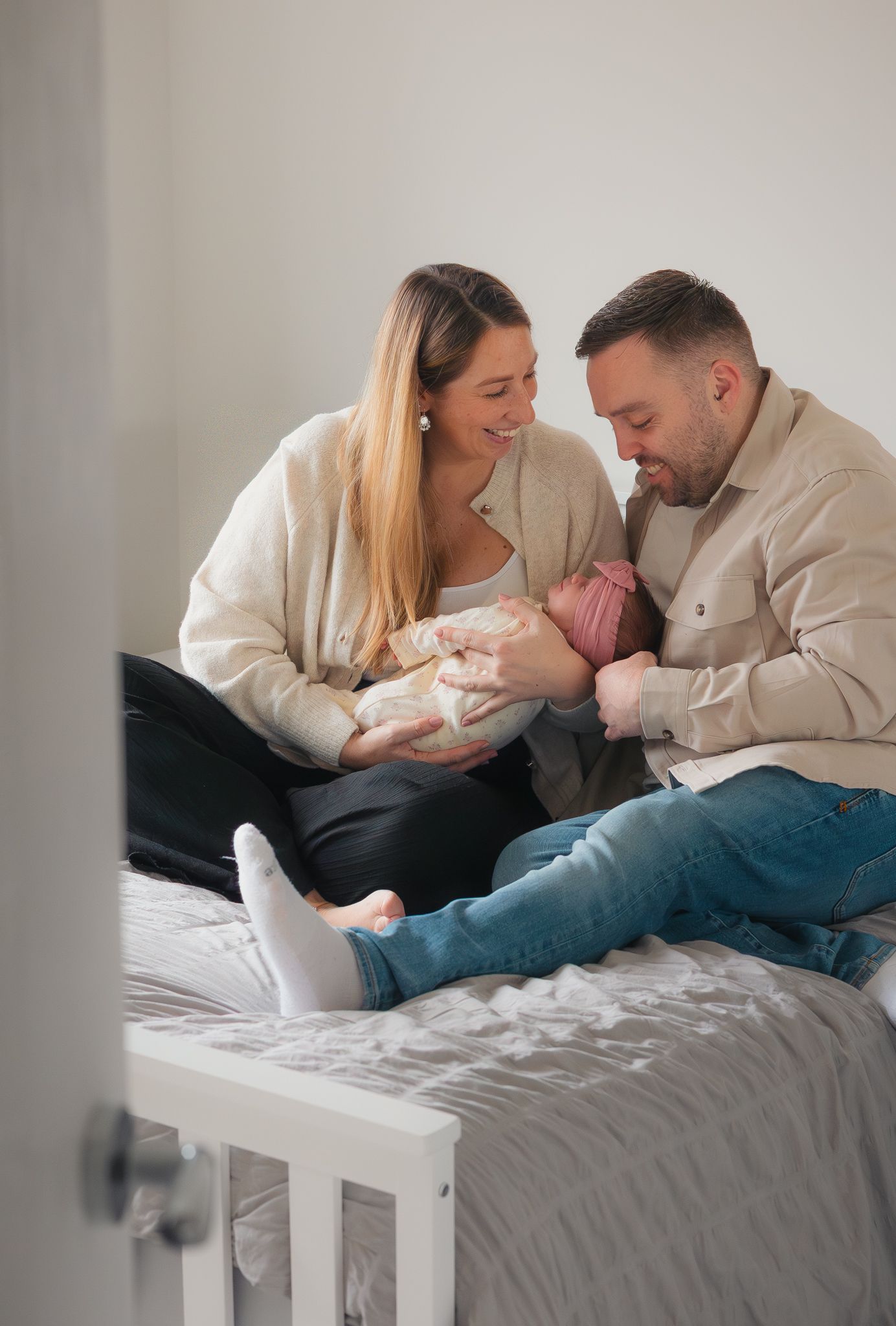 A happy couple sitting on a bed, holding a newborn baby and smiling affectionately in Sevenoakes.