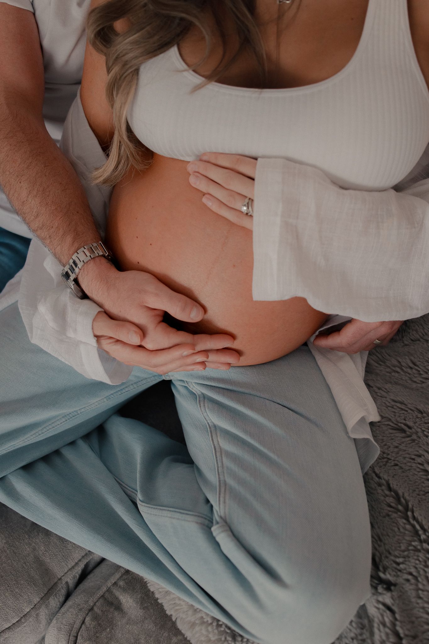Couple cradling pregnant belly together, man wearing watch placing hand over woman's baby bump during maternity photo session in Cricklade.