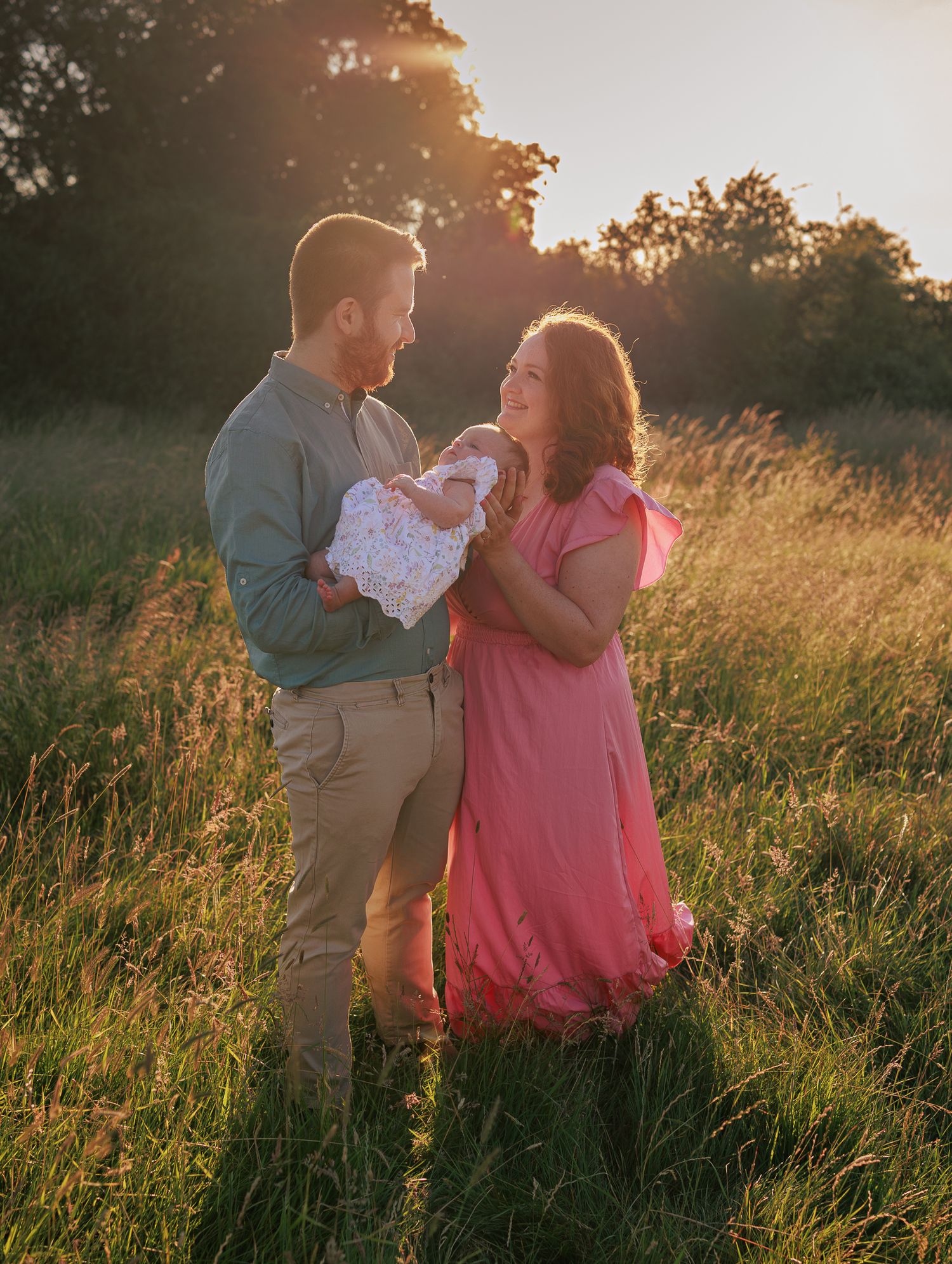 A couple stands in a sunlit meadow at golden hour, the man holding a newborn baby as the woman in a pink dress smiles warmly in New Forest.