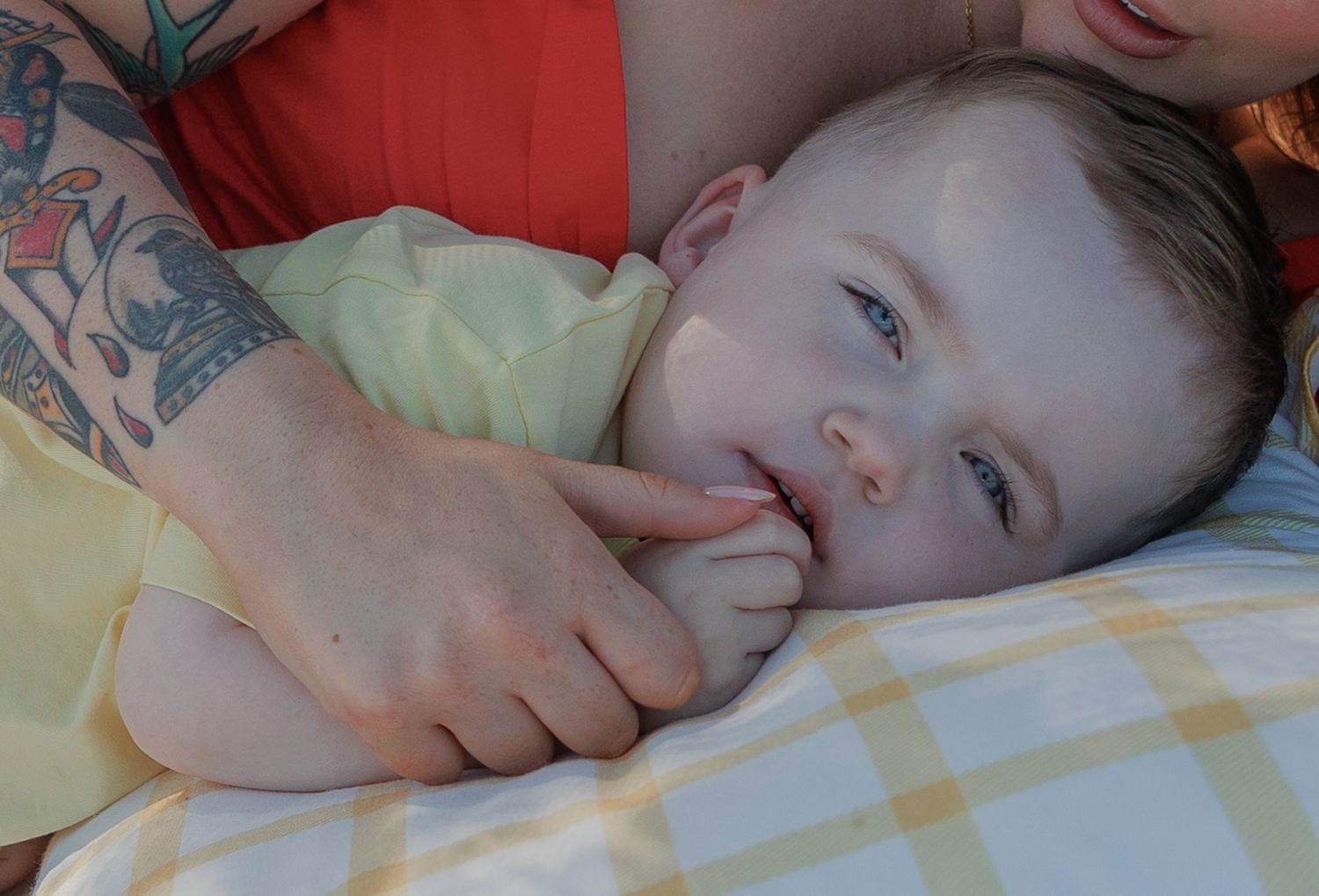 A baby with blue eyes lies on a yellow blanket, gently held by a tattooed adult's hand in Fairford, Cotswolds.