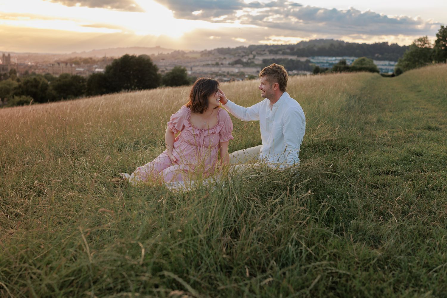 Couple sitting in golden hour sunlit meadow, for a Maternity Photoshoot in Cotswolds, UK.