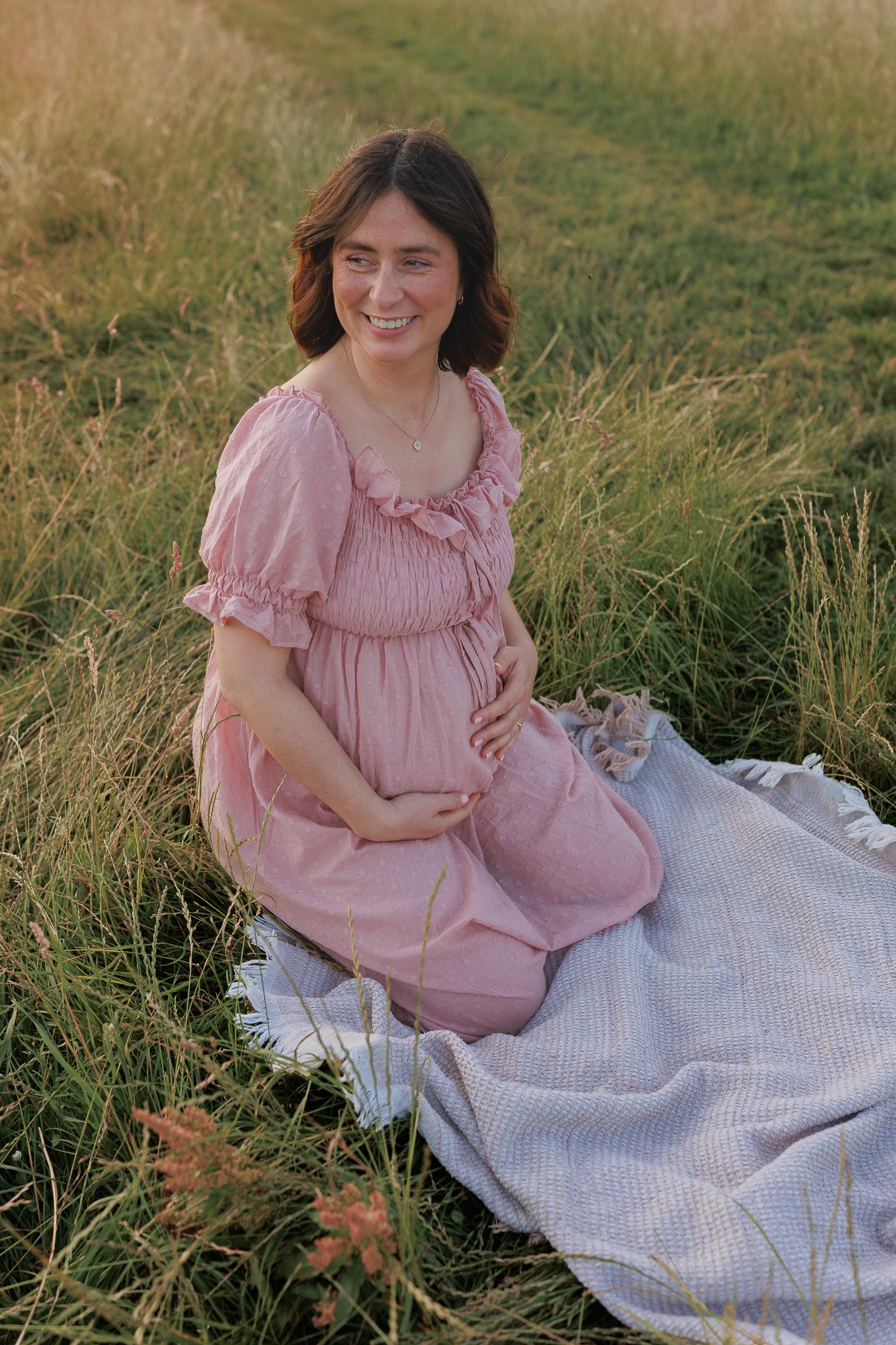 Smiling pregnant woman in pink dress sits on blanket in golden grass field at sunset in Berkshire.